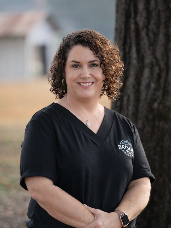 A woman with curly brown hair smiling outdoors, wearing a black shirt with a logo, standing near a tree.