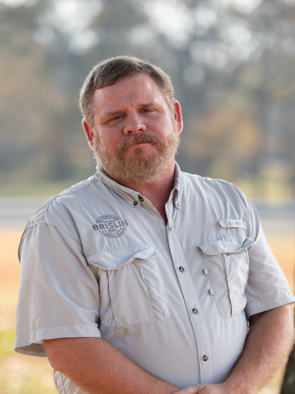 A man with a beard and mustache wearing a light grey short-sleeved button-up shirt outdoors.
