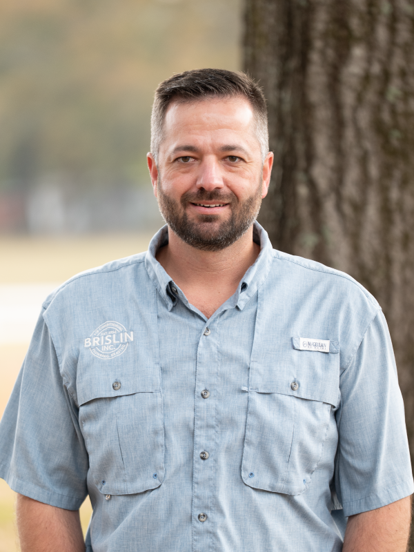 A man with short dark hair, a beard, and a light blue uniform shirt standing outdoors near a large tree with blurred background. The shirt has a logo and name tag.