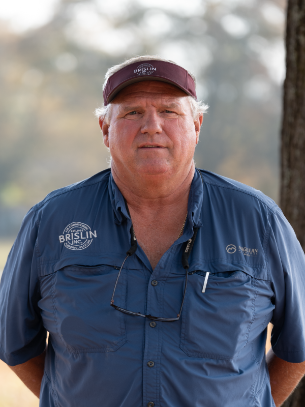 A middle-aged man with white hair wearing a maroon visor, blue outdoor shirt with 'Brislin' logo, and sunglasses hanging around his neck. He is standing outdoors with trees in the background.