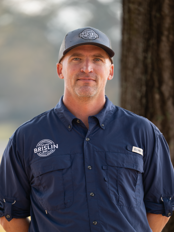 A man wearing a blue work shirt with company logos and a gray cap standing outdoors near a tree with blurred background.