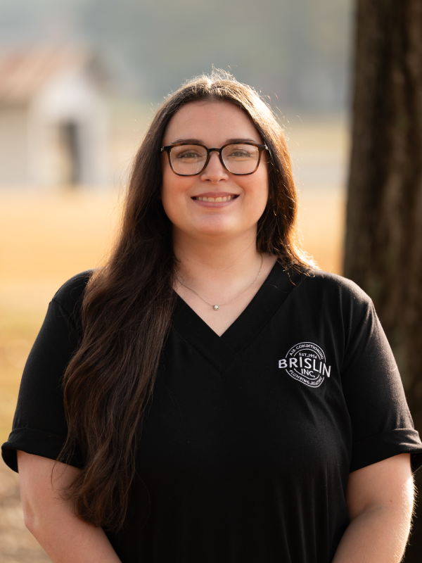 A woman with long dark hair and glasses smiling outdoors, wearing a black polo shirt with a logo.