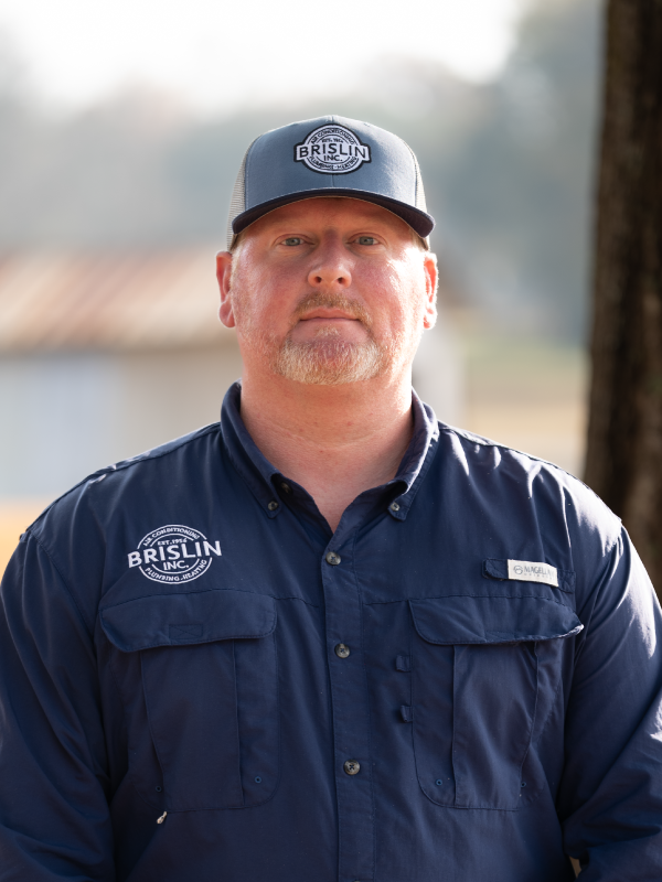 Man with light skin, reddish beard, wearing a blue work shirt and a gray trucker hat with a logo, standing outdoors with blurred background.