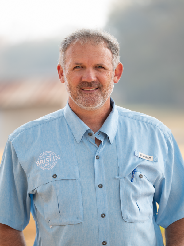 A middle-aged man with gray hair and beard, wearing a light blue work shirt with company logos, standing outdoors with a blurred landscape background.