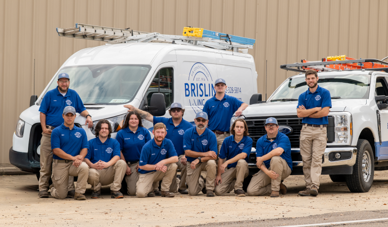 A team of eleven uniformed Brislin, Inc. workers in blue shirts and tan pants standing and kneeling in front of service trucks, posing outdoors in front of a beige building.