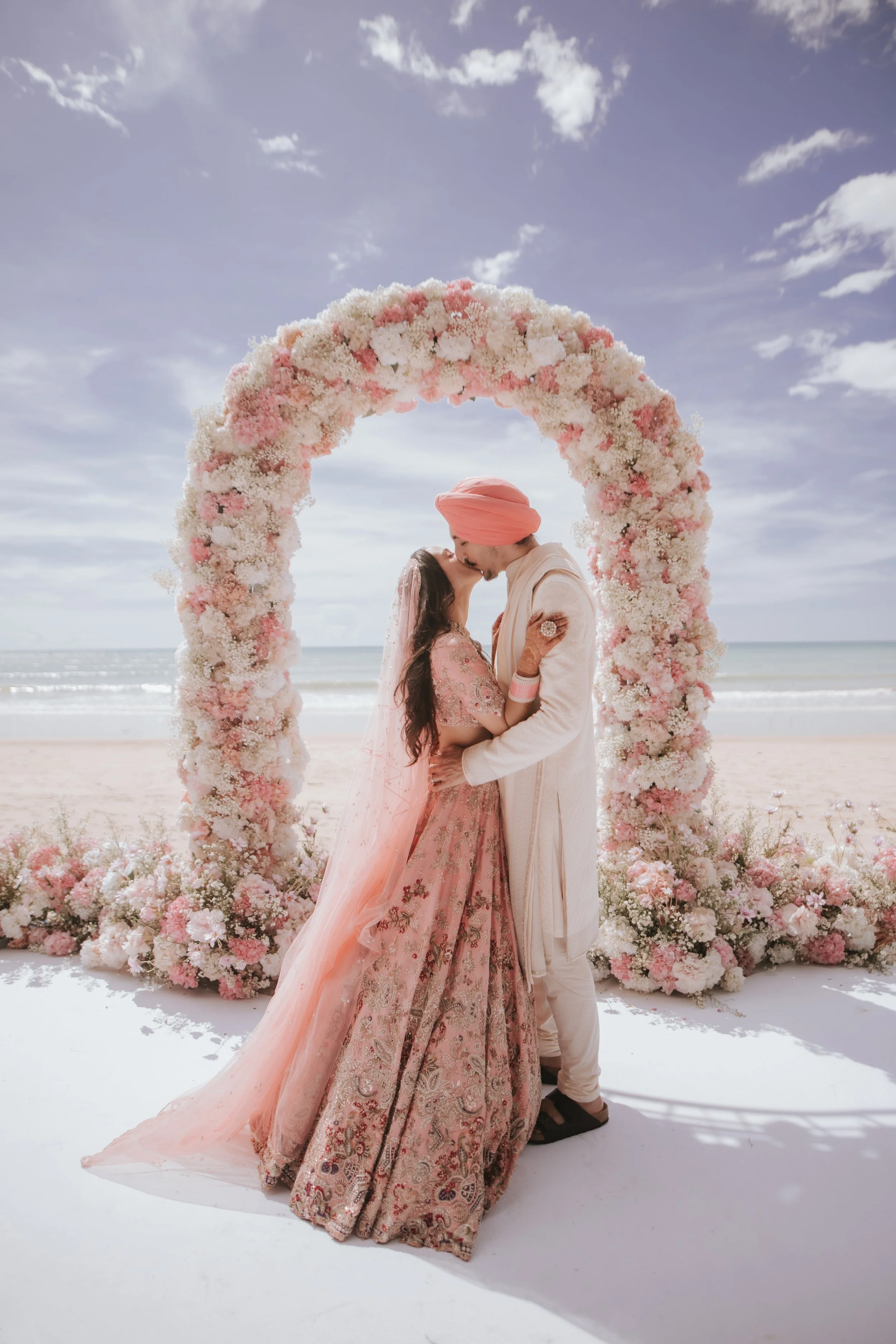 A couple in traditional Indian wedding attire sharing a kiss under a floral arch on a beach.