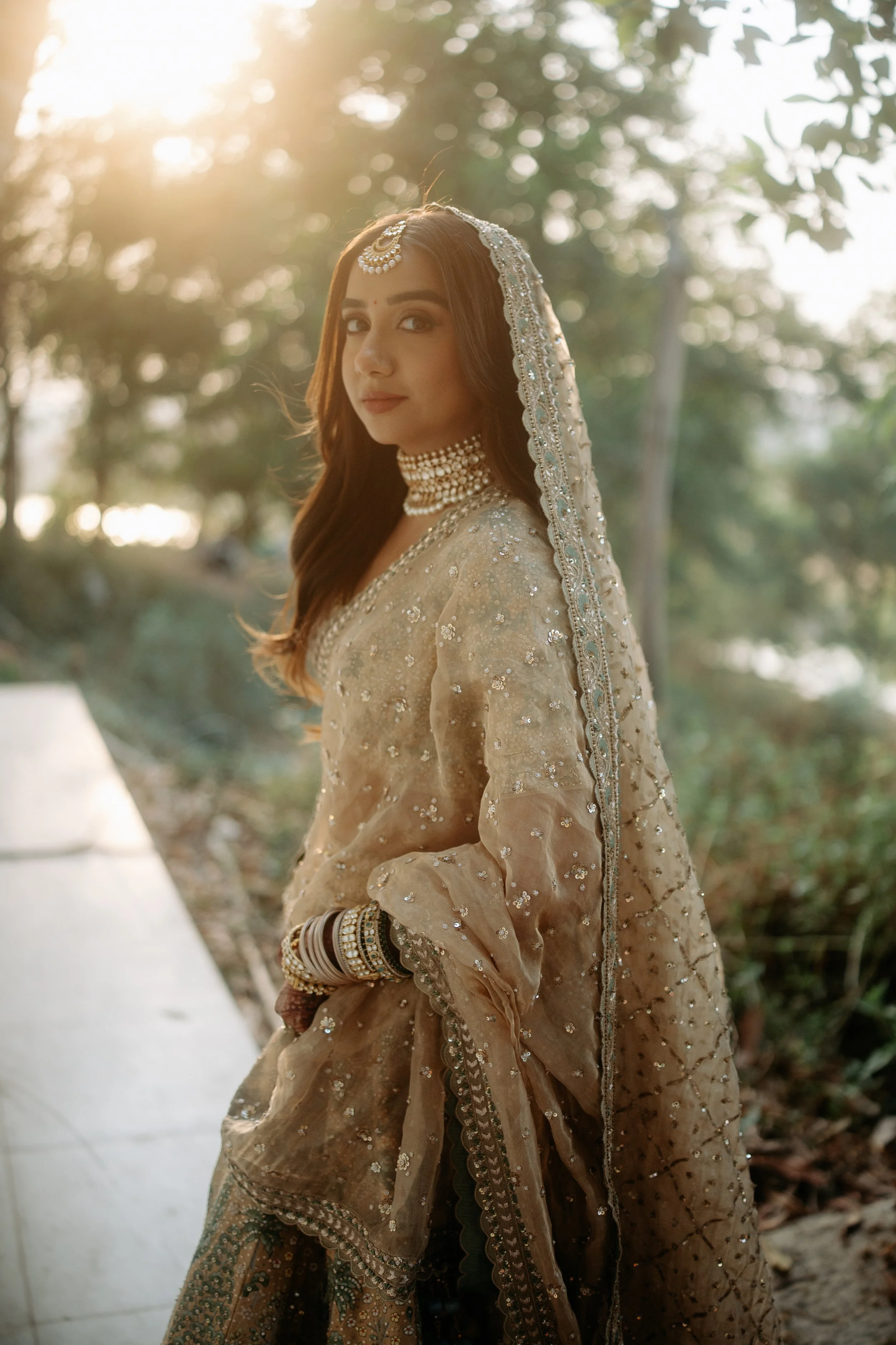 A woman dressed in traditional Indian bridal attire, wearing a gold and silver embroidered lehenga with matching jewelry, standing outdoors with sunlight filtering through trees.