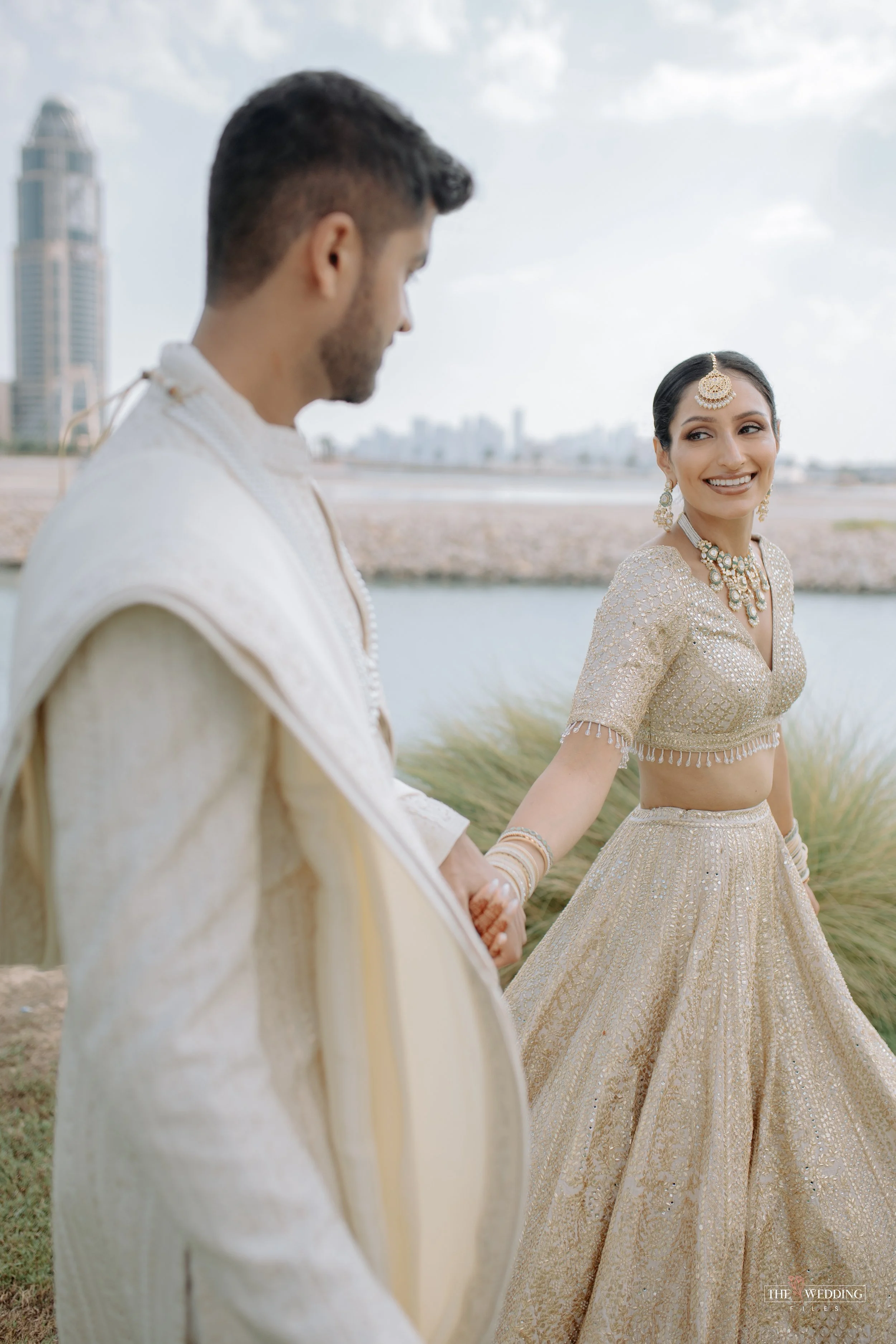A bride in a traditional Indian gold and cream lehenga holding hands with a groom in a cream sherwani, outdoors near a water body with city buildings in the background.