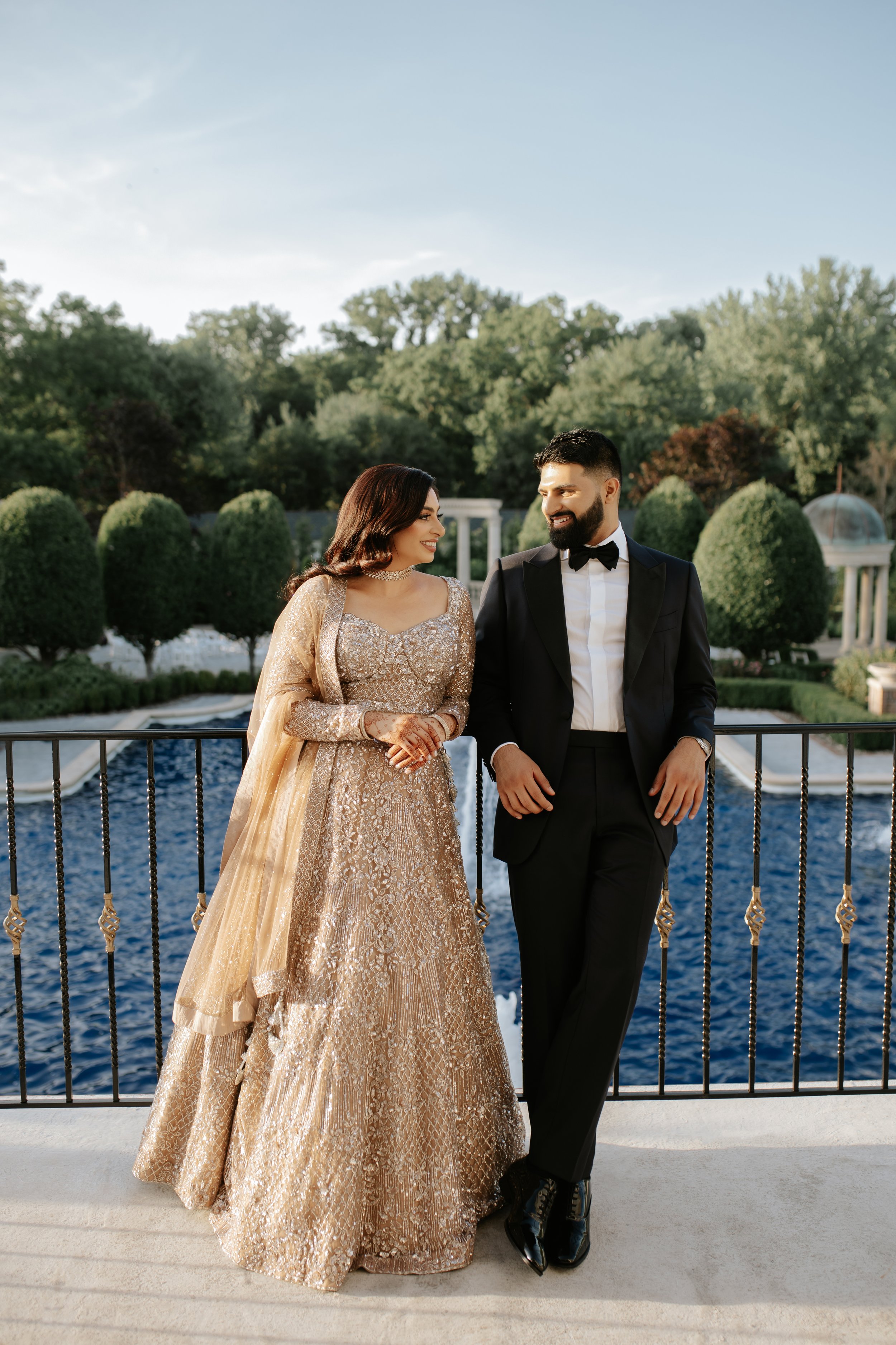 A couple dressed in formal attire standing near a balcony railing overlooking a pool and garden with trees in the background.