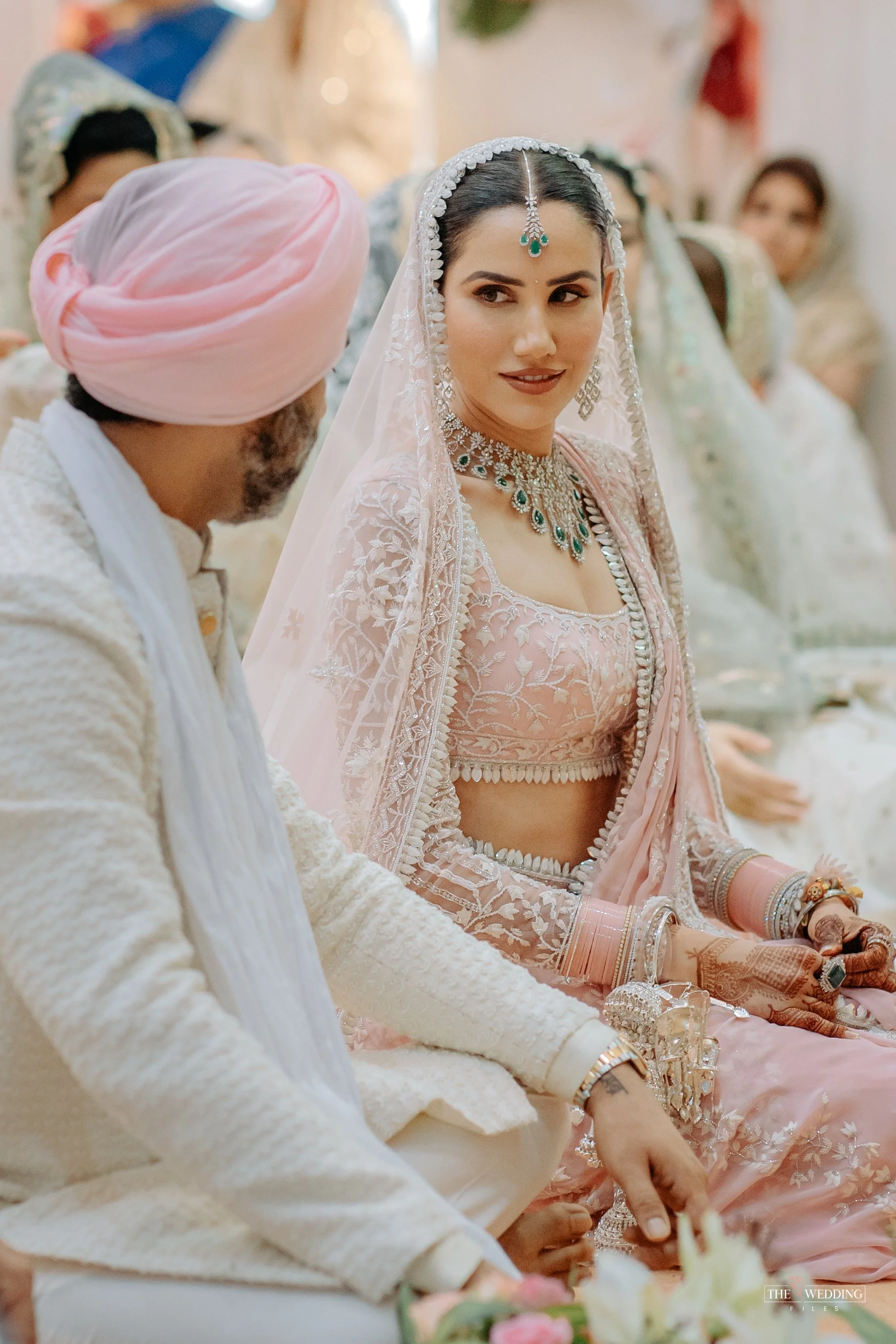 A bride in a pink traditional wedding dress with jewelry, sitting next to a man in a cream-colored outfit with a pink turban, during a wedding ceremony. Other guests are visible in the background.