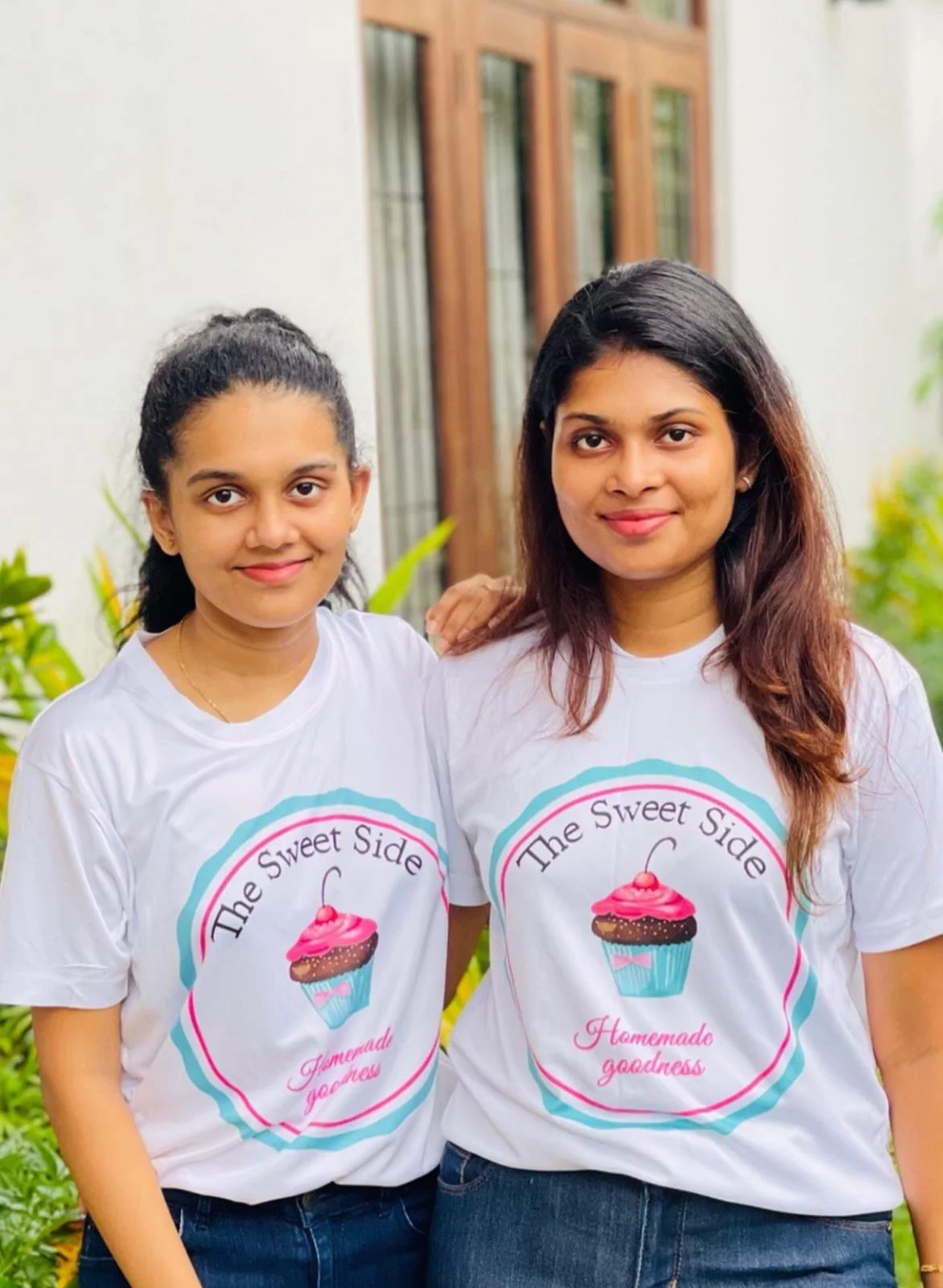 Two women standing outside, wearing matching white t-shirts with a cupcake graphic and the text 'The Sweet Side Homemade goodness.'