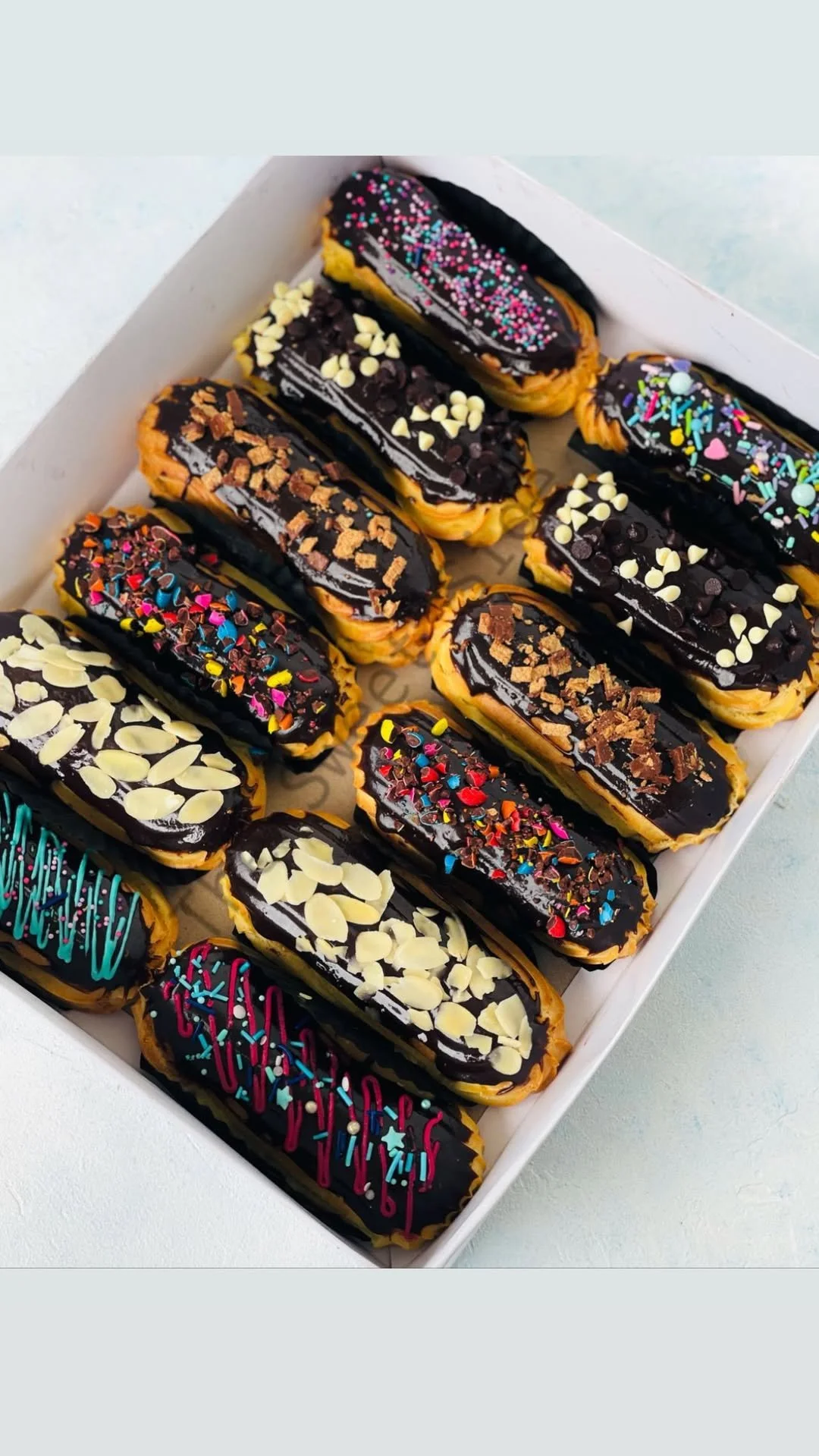 Assorted éclairs topped with chocolate glaze and colorful sprinkles or nuts in a white box.