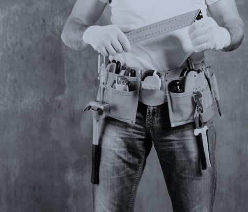 Person wearing work utility belt filled with hand tools, holding a ruler, with a plain background.