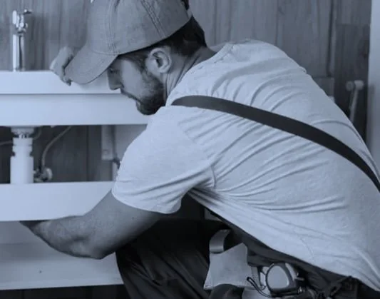 A man wearing a cap and T-shirt working under a kitchen sink.