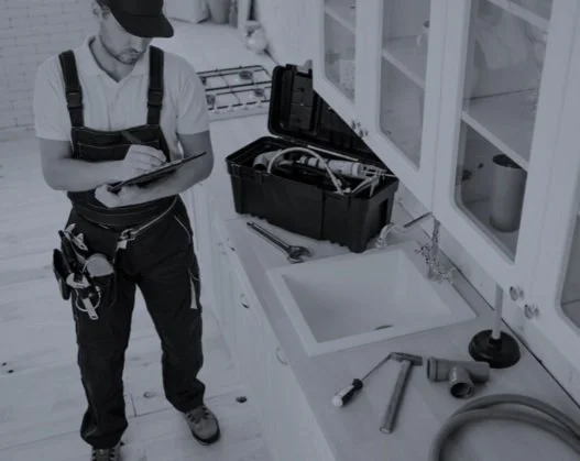 A plumber wearing a cap and work overalls standing at a kitchen sink, holding a tablet, with plumbing tools and a toolbox nearby.