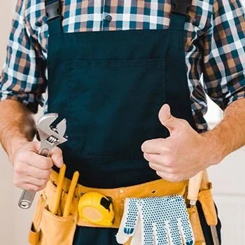 Person wearing a plaid shirt and black apron, holding a wrench with a tool belt that contains various tools including a tape measure, gloves, and screwdrivers.