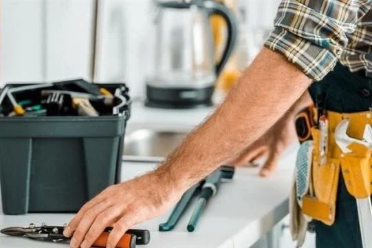 Close-up of a person's arm wearing a checkered shirt, with a tool belt, working at a kitchen countertop with tools and a toolbox.