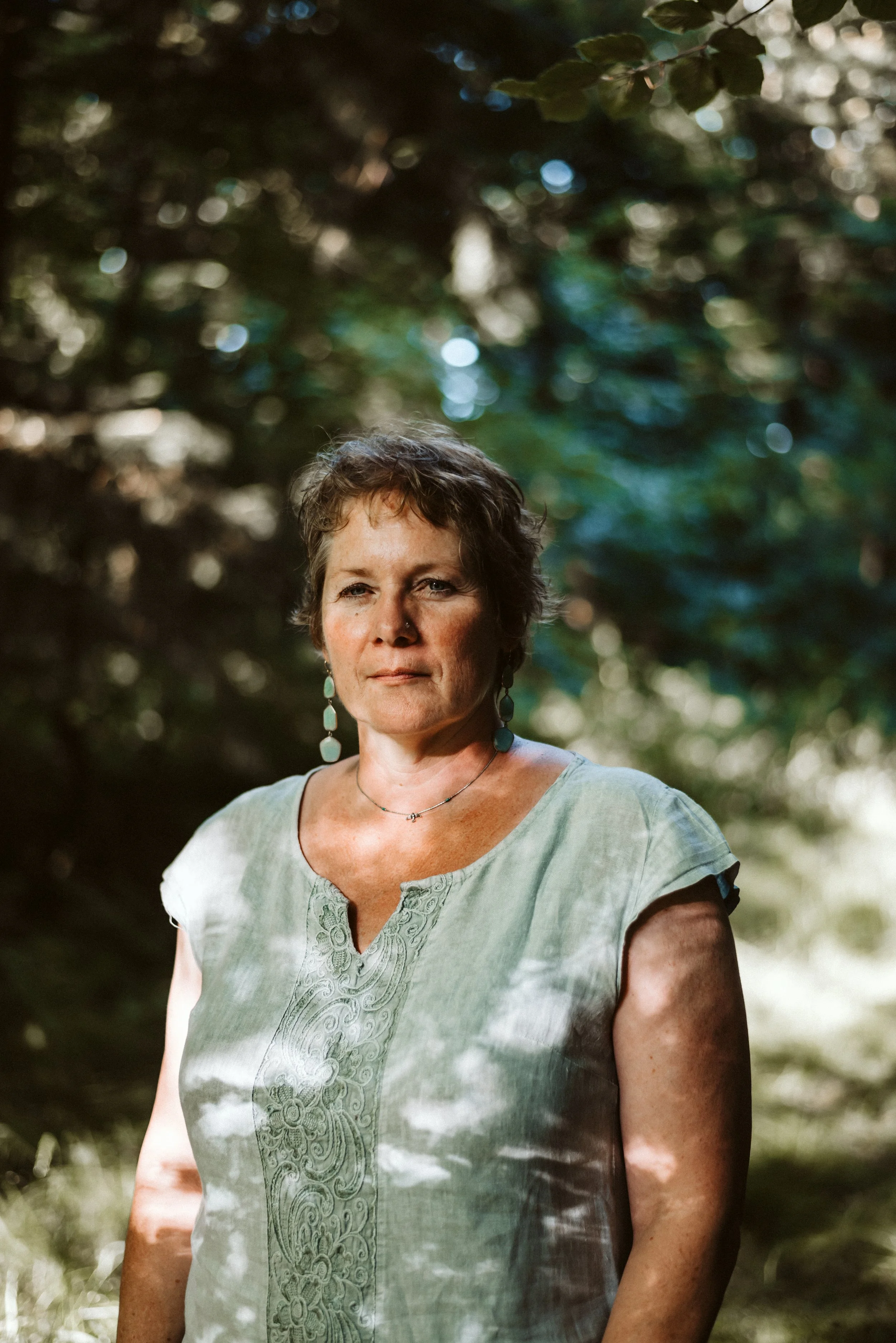 Eine Frau steht in einem Wald, umgeben von Bäumen und Sonnenlicht, das durch die Blätter scheint.