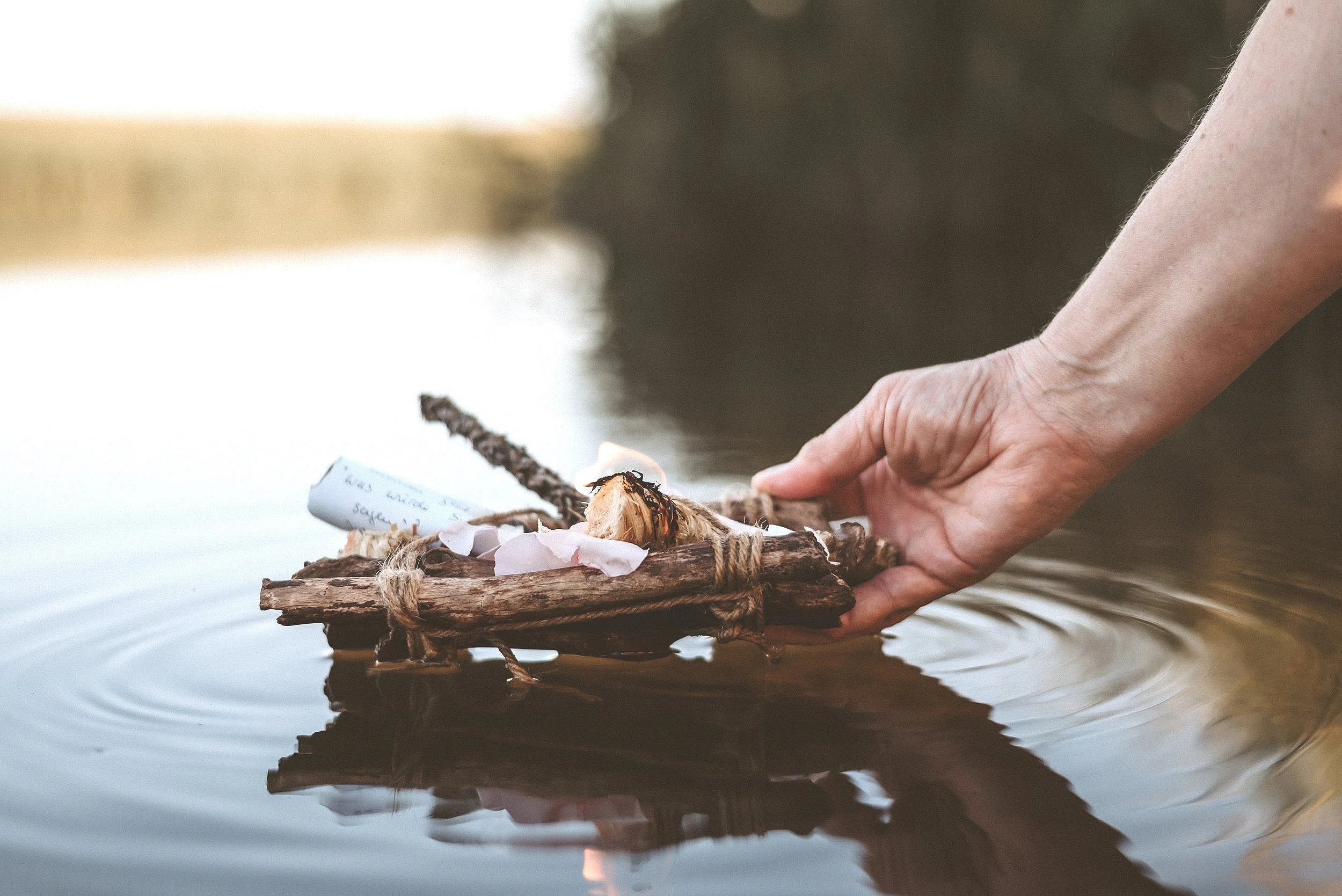 Eine Hand hält ein Floß aus Holzstücken auf einem Wasserteich, auf dem ein Zettel und Blüten liegen.