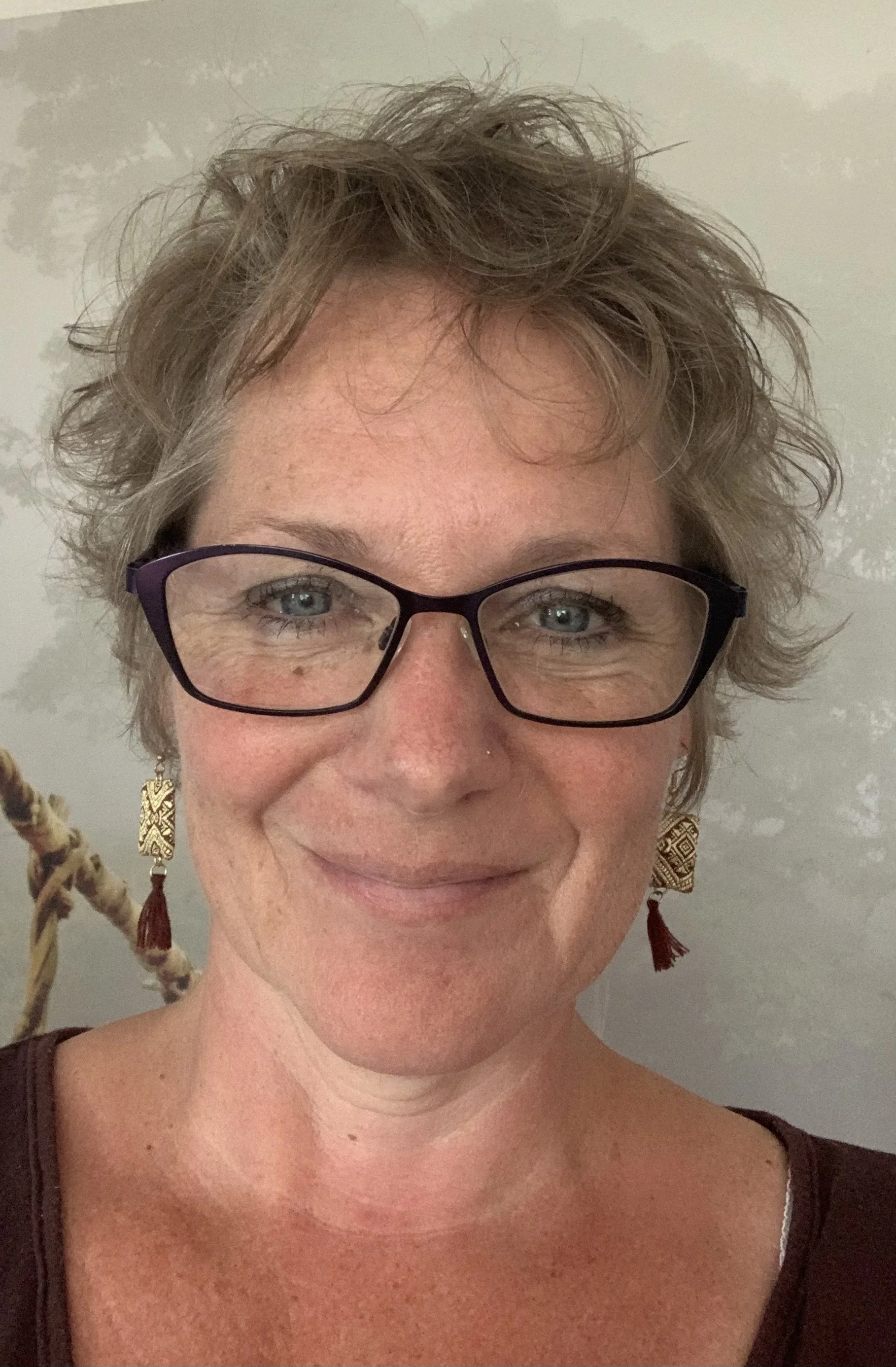 Nahaufnahme eines lachenden Frauenkopfes mit kurzen, lockigen Haaren, Brille und Ohrringen vor einem neutralen Hintergrund.