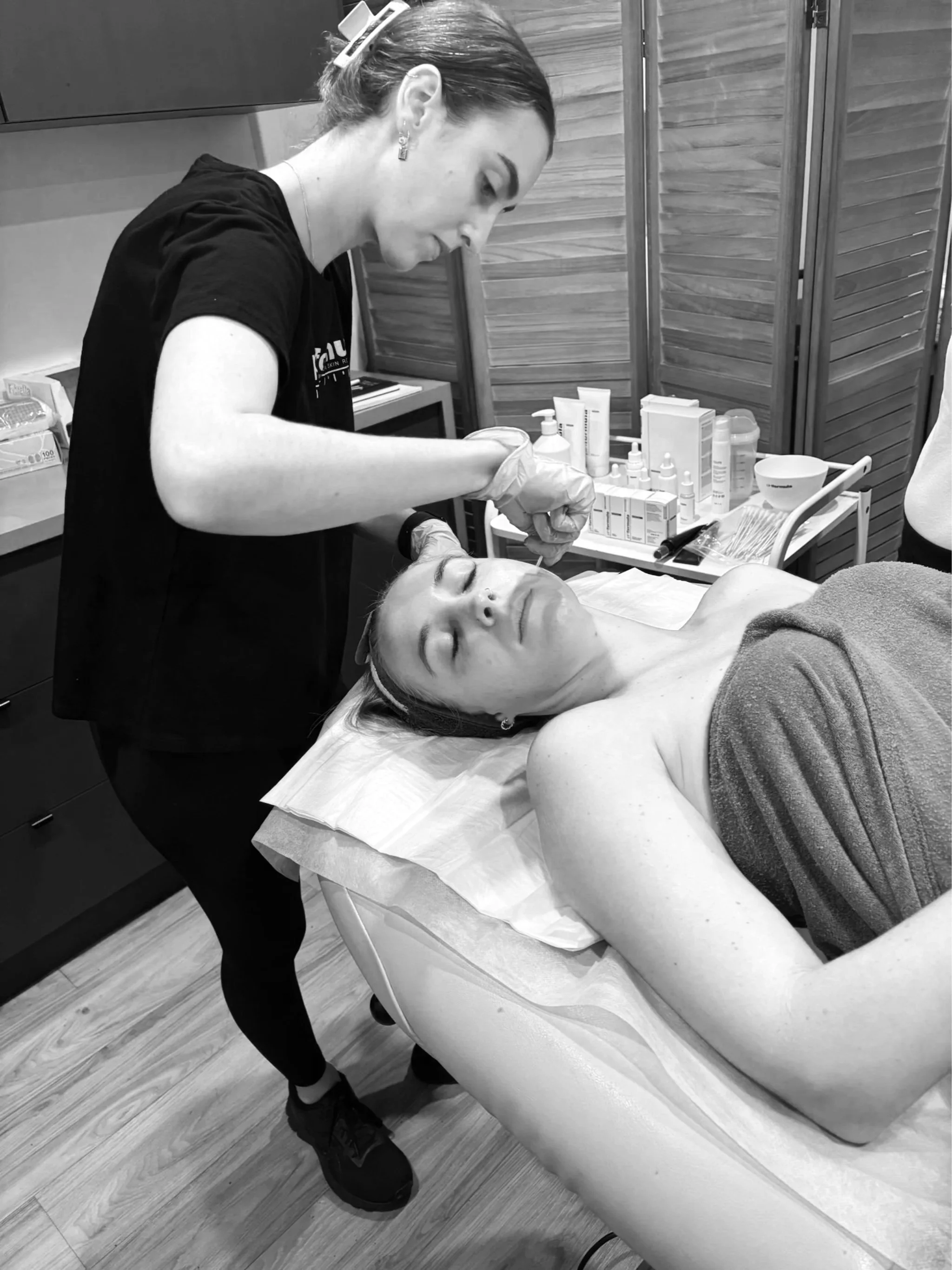 A woman lying on a treatment bed with her eyes closed, receiving a cosmetic or skincare treatment from a practitioner in a clinic or spa setting.