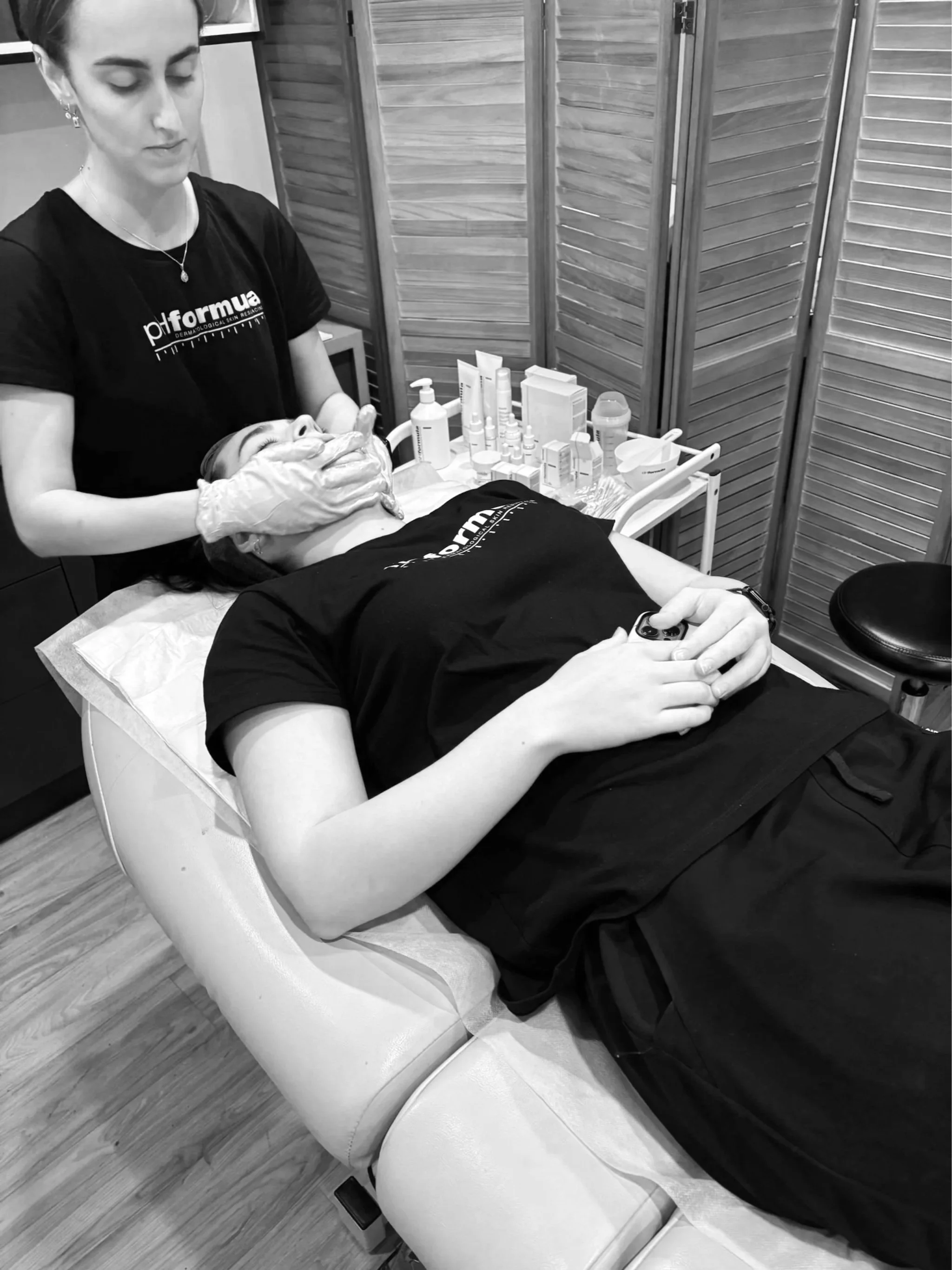 A woman lying on a treatment bed with her eyes closed, receiving a facial treatment from a skincare professional in a clinical setting.