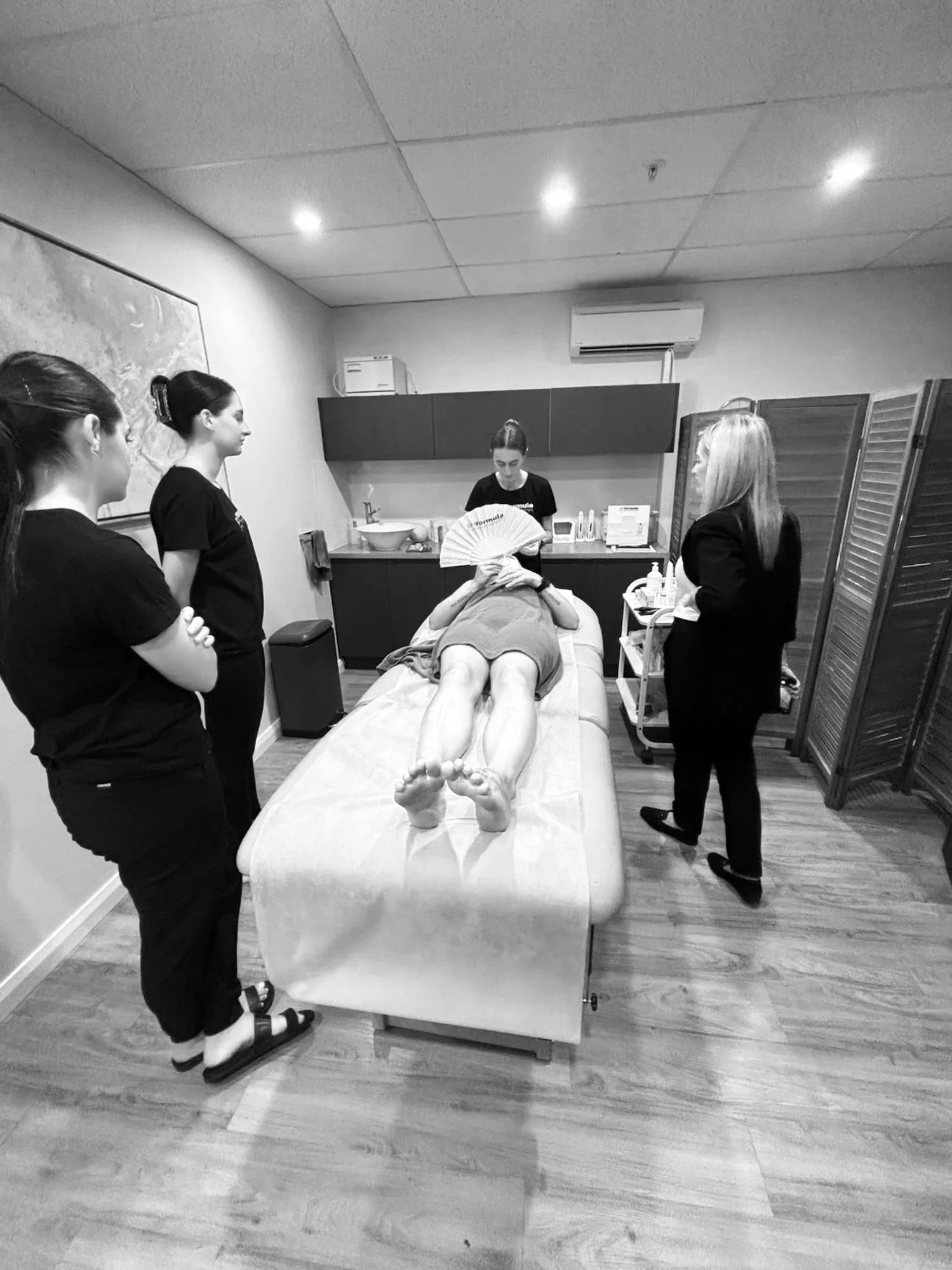 A woman lying on a massage table with a fan covering her face, surrounded by four women and one man in a professional setting, possibly a spa or clinic.