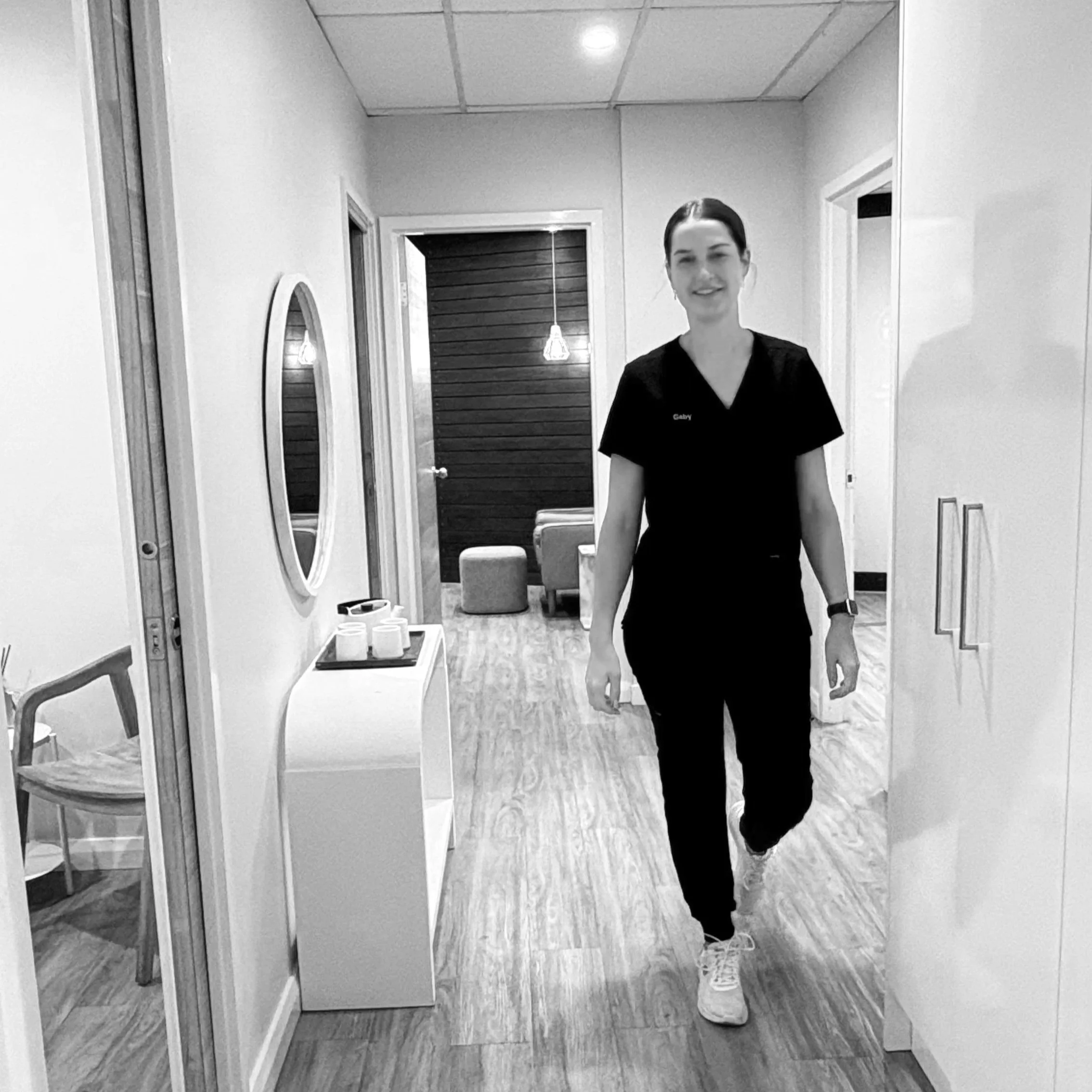 A woman in medical scrubs walking down a hallway in a modern lounge or office space, smiling at the camera.