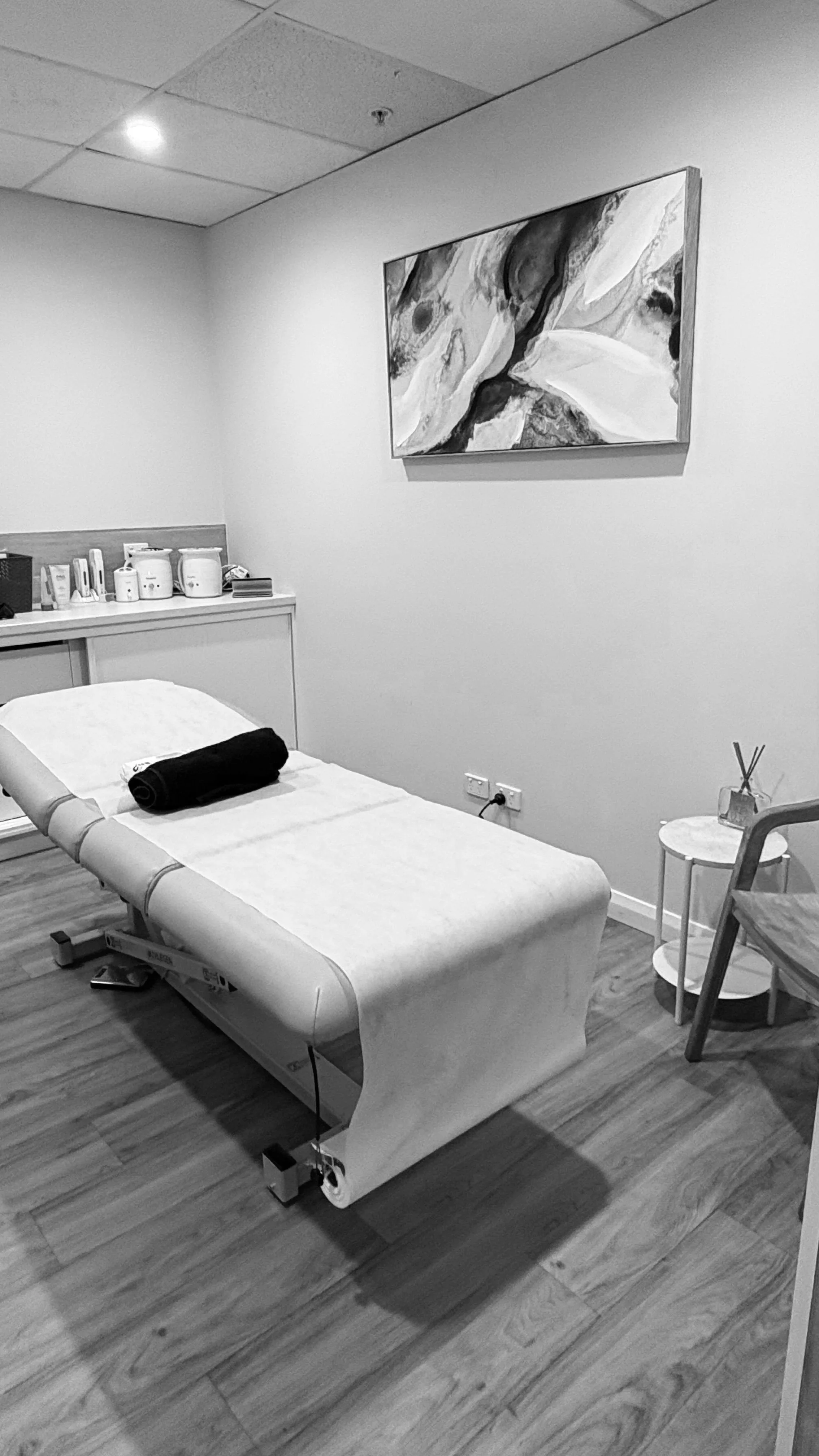 An empty examination or massage room with a treatment bed covered with a towel and a rolled black blanket, a small side table, abstract painting on the wall, and medical supplies on a cabinet.