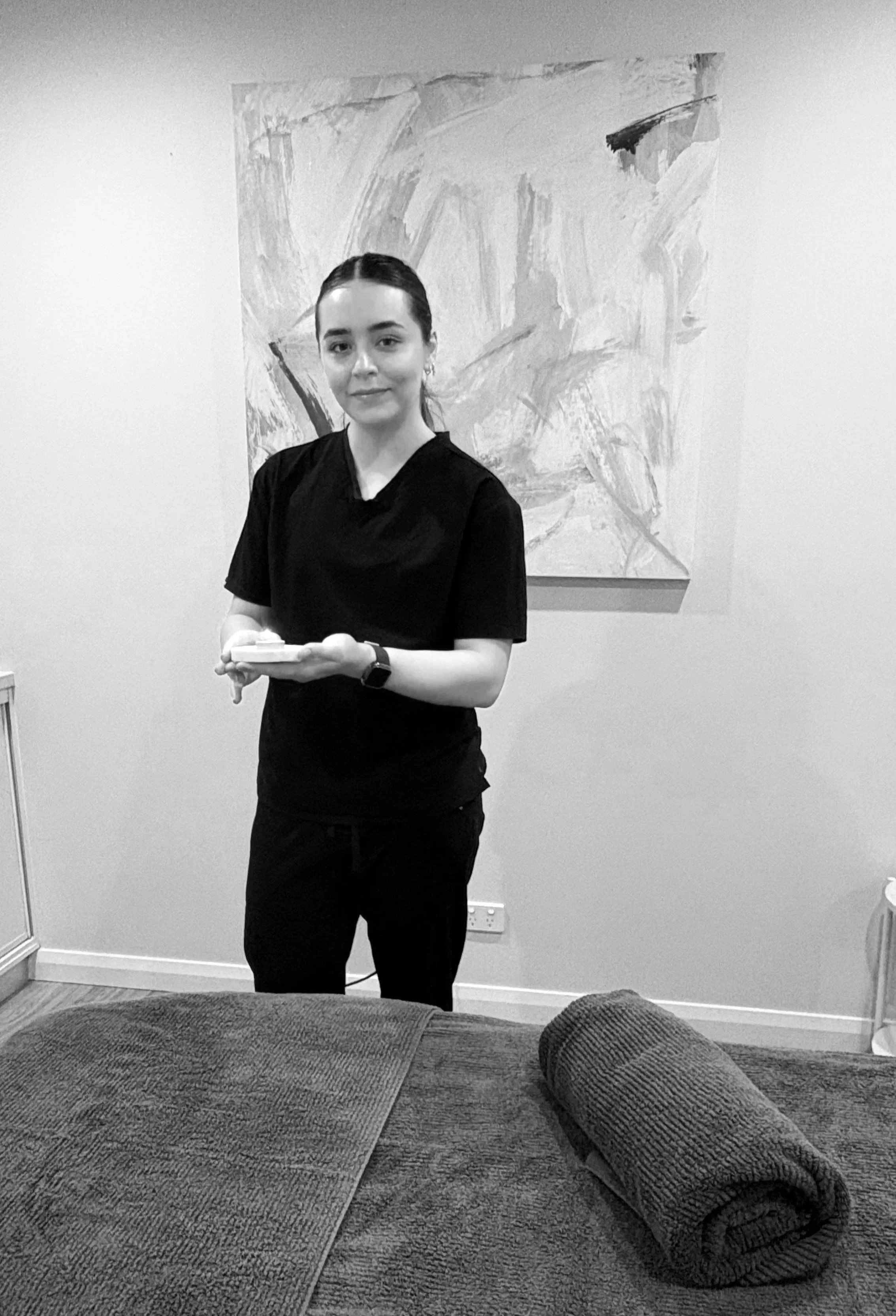 A woman in black scrubs standing in a room, holding a small tray, with a rolled towel on a massage table in the foreground and an abstract painting on the wall behind her.