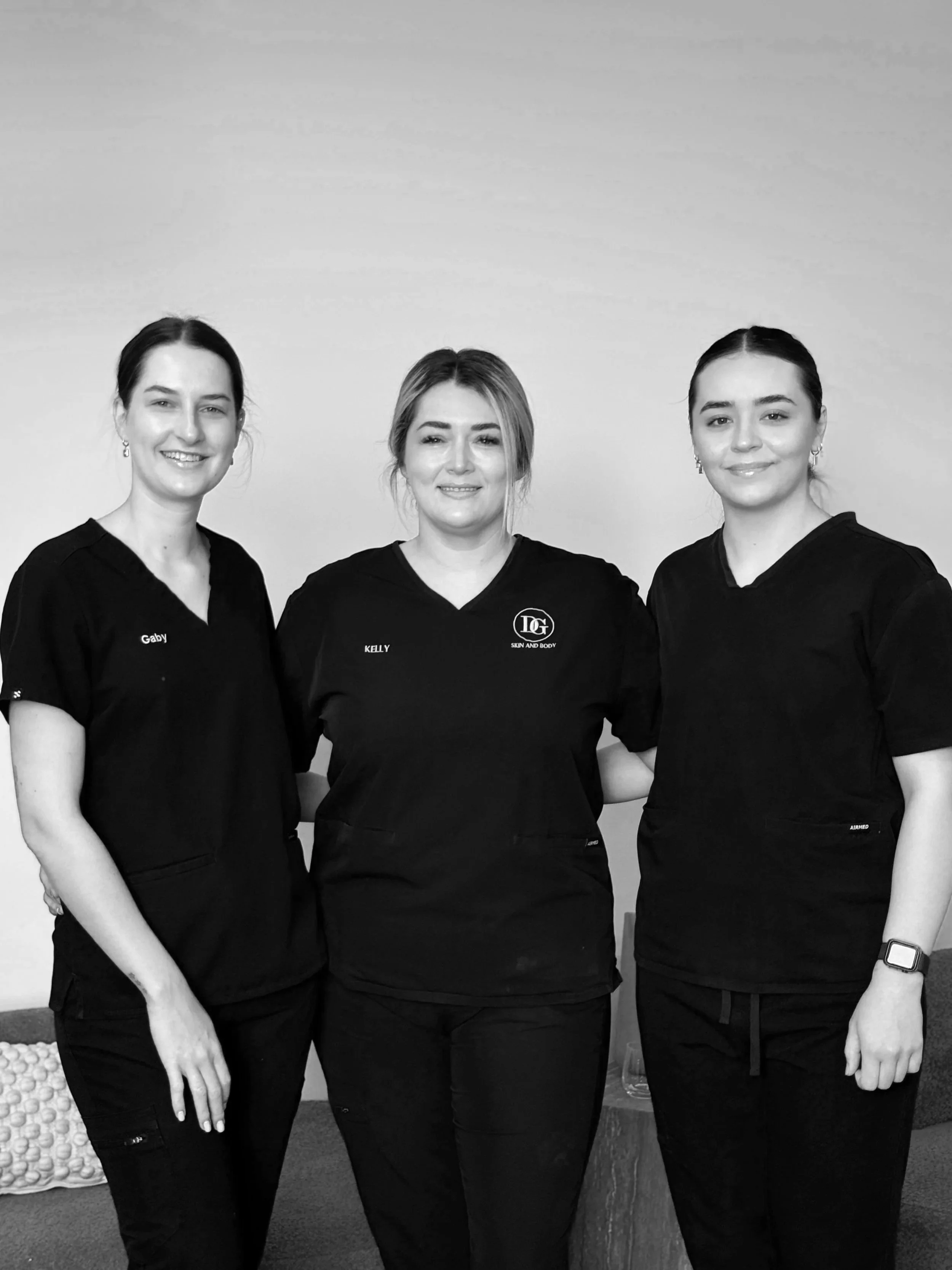 Three women in black medical scrubs standing together in front of a plain wall, smiling at the camera.
