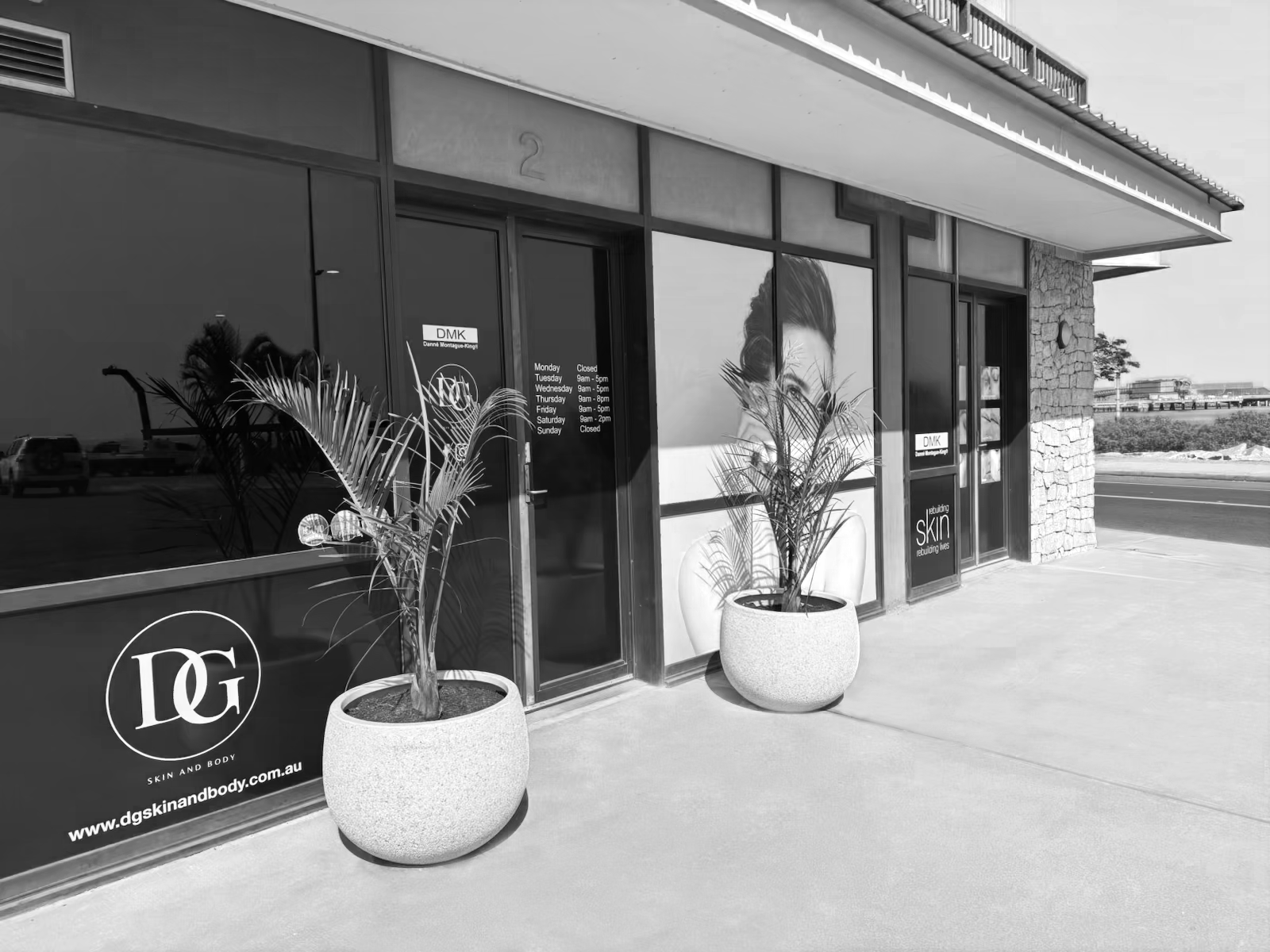 Exterior of a skincare clinic with large glass windows, two potted plants outside, and promotional images for skin treatments. Business hours are posted on the door.