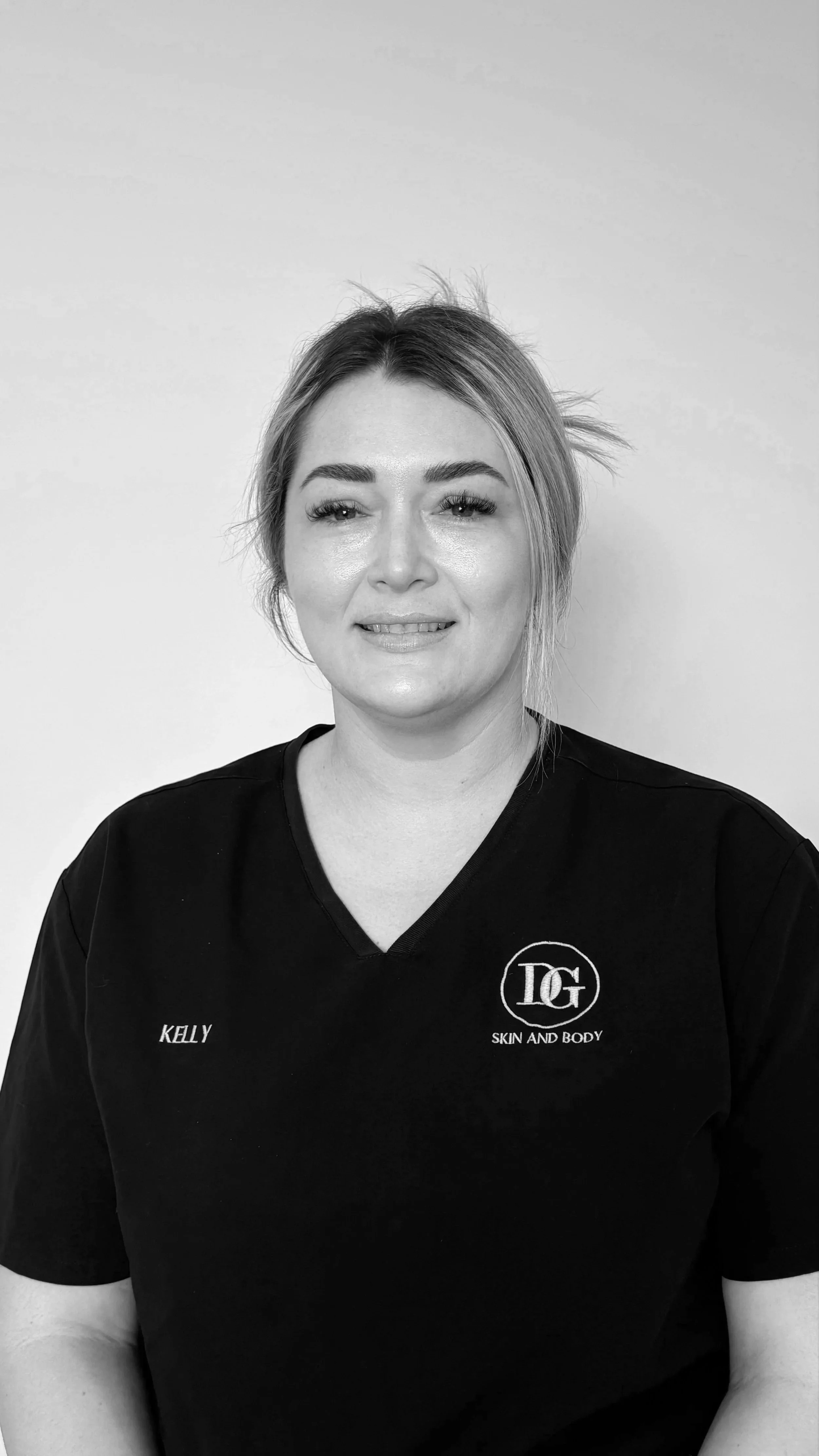 Black and white portrait of a woman with light hair tied back, wearing a black t-shirt with the logo 'DG' and the text 'Skin and Body', and the name 'Kelly' embroidered on her shirt.