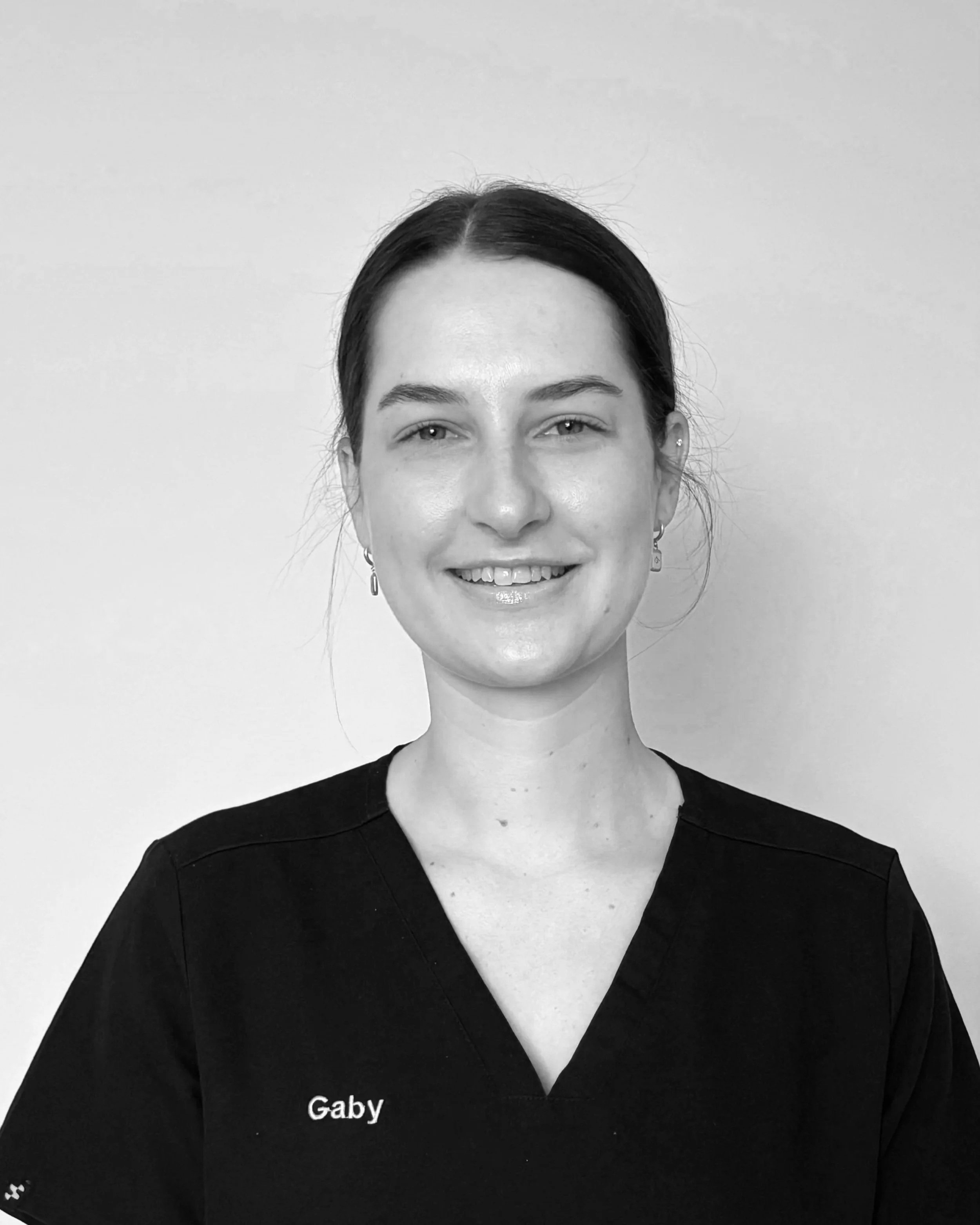 Black and white portrait of a smiling woman with dark hair pulled back, wearing earrings, a dark V-neck top with 'Gaby' embroidered on it, standing against a plain background.