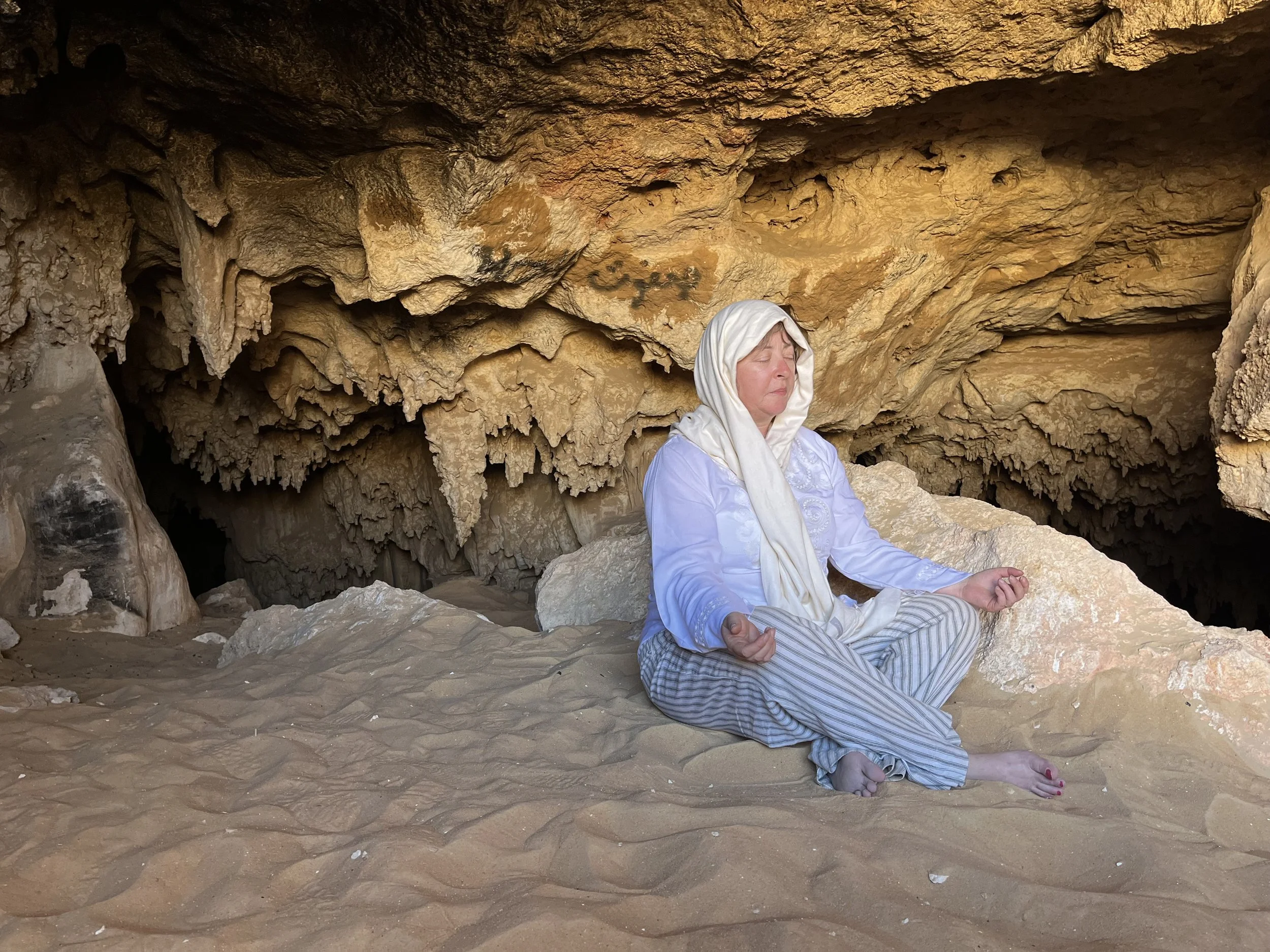 A woman dressed in white and gray striped pants sits cross-legged on sandy ground inside a cave, meditating with her eyes closed and wearing a white headscarf.