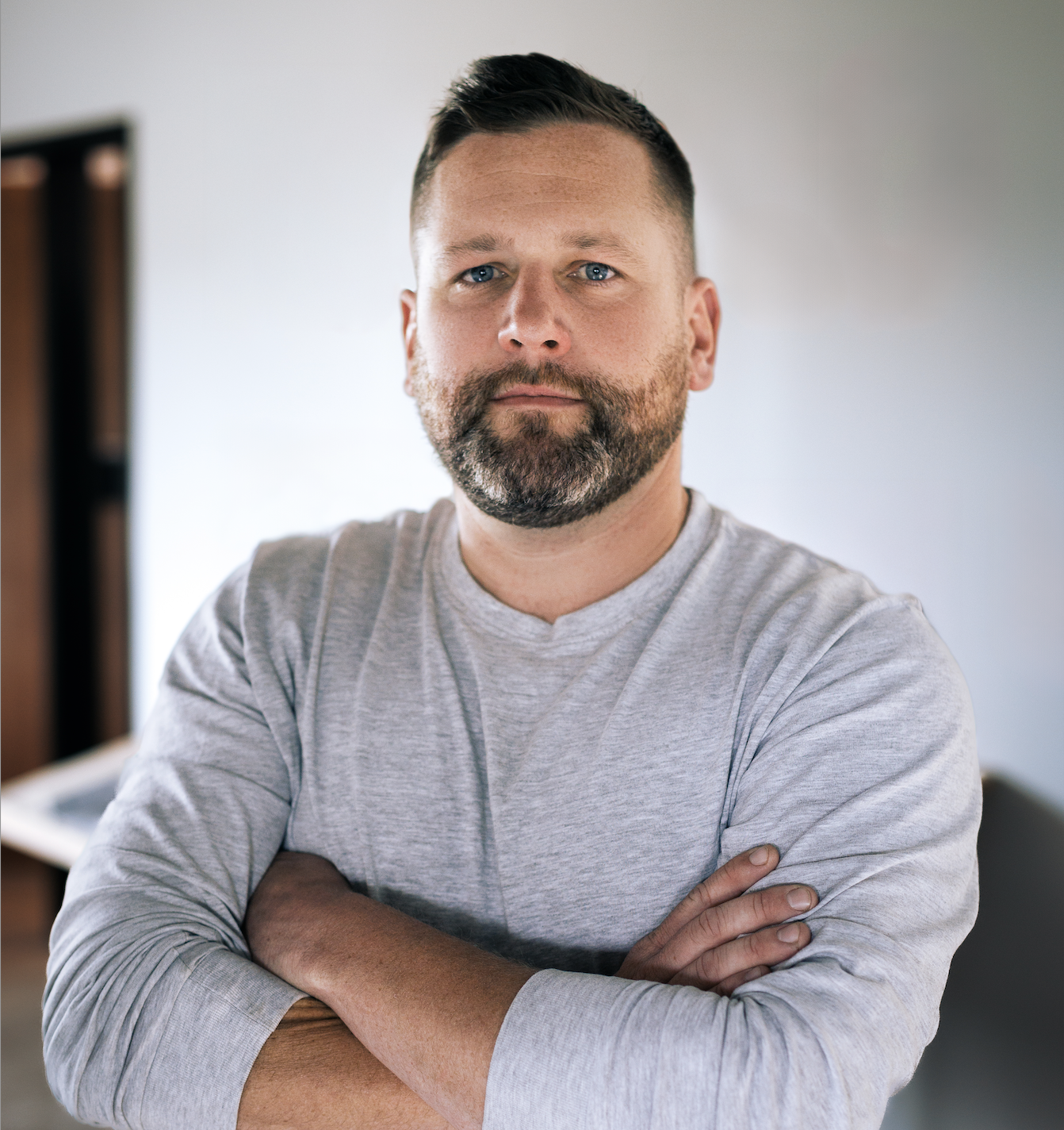 A smiling man with a beard and short dark hair, wearing a gray long-sleeve shirt, standing with arms crossed in a room with neutral-colored walls.