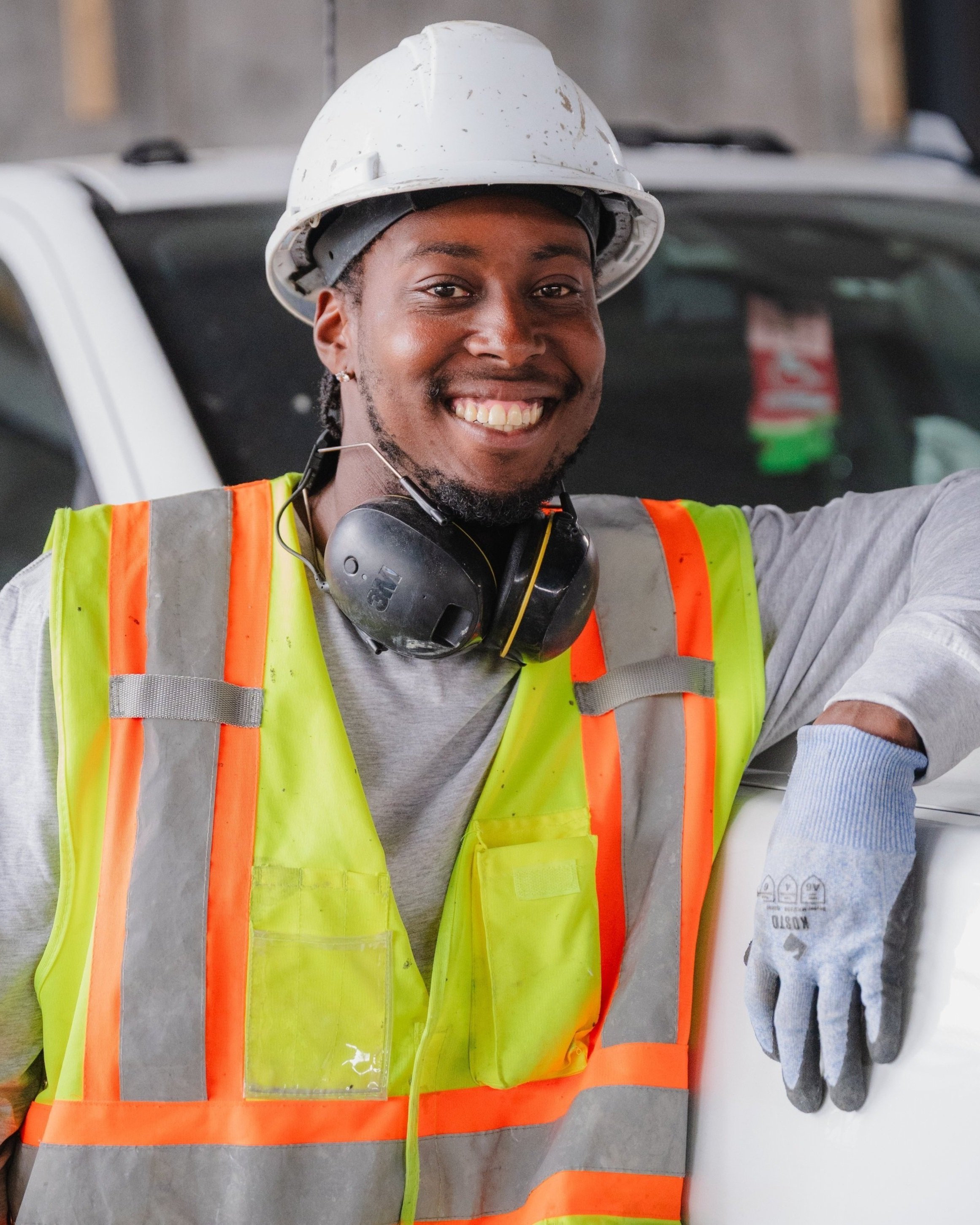 A smiling construction worker wearing a white hard hat, safety vest, gloves, and ear protection, standing beside a vehicle.