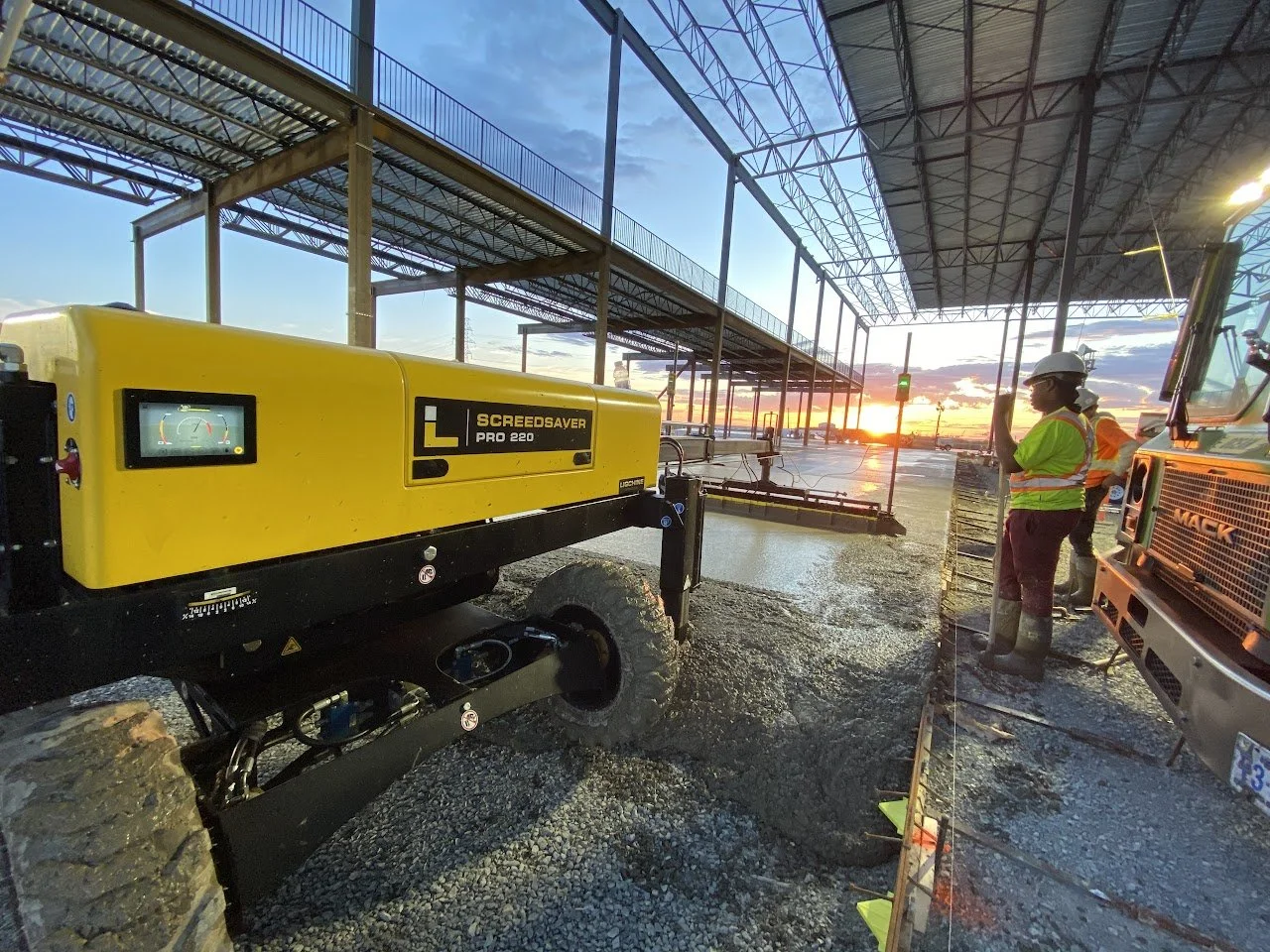 Construction workers wearing safety gear working on a building site during sunset, with heavy machinery and partially completed structure.