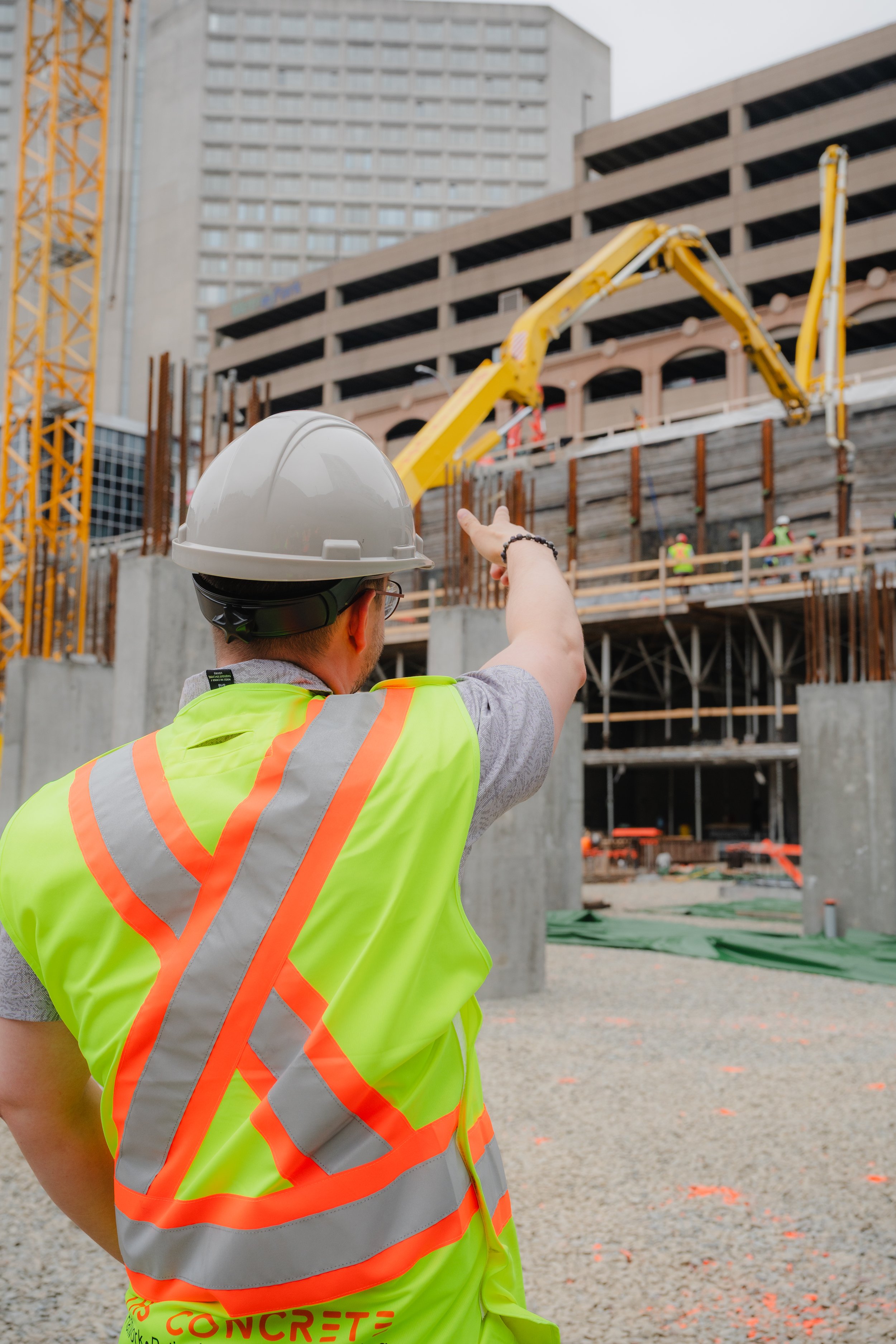 Construction worker in a yellow safety vest and hard hat pointing at a building under construction with cranes and scaffolding.