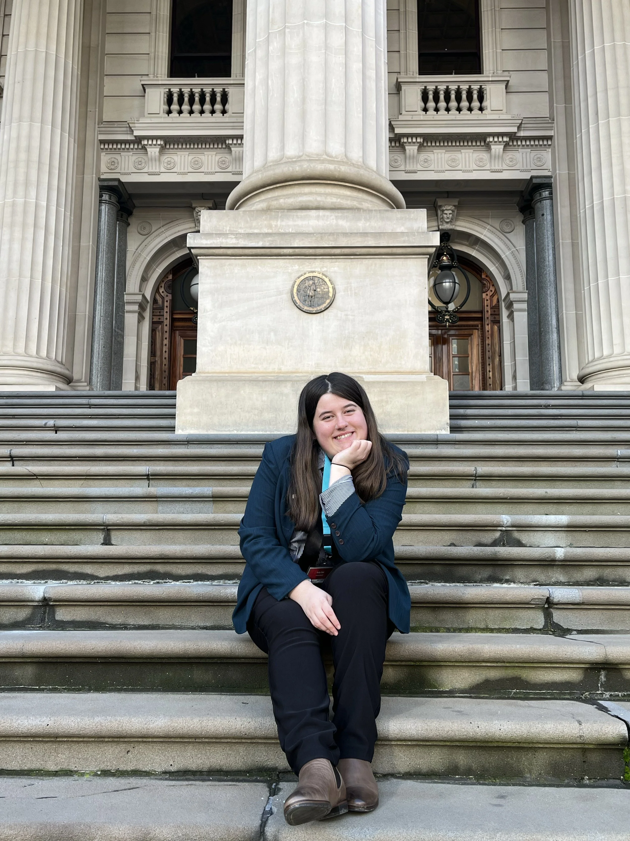 A young woman with long brown hair sitting on stone steps outside a grand government building, smiling at the camera, wearing a dark blazer, black pants, and brown boots.