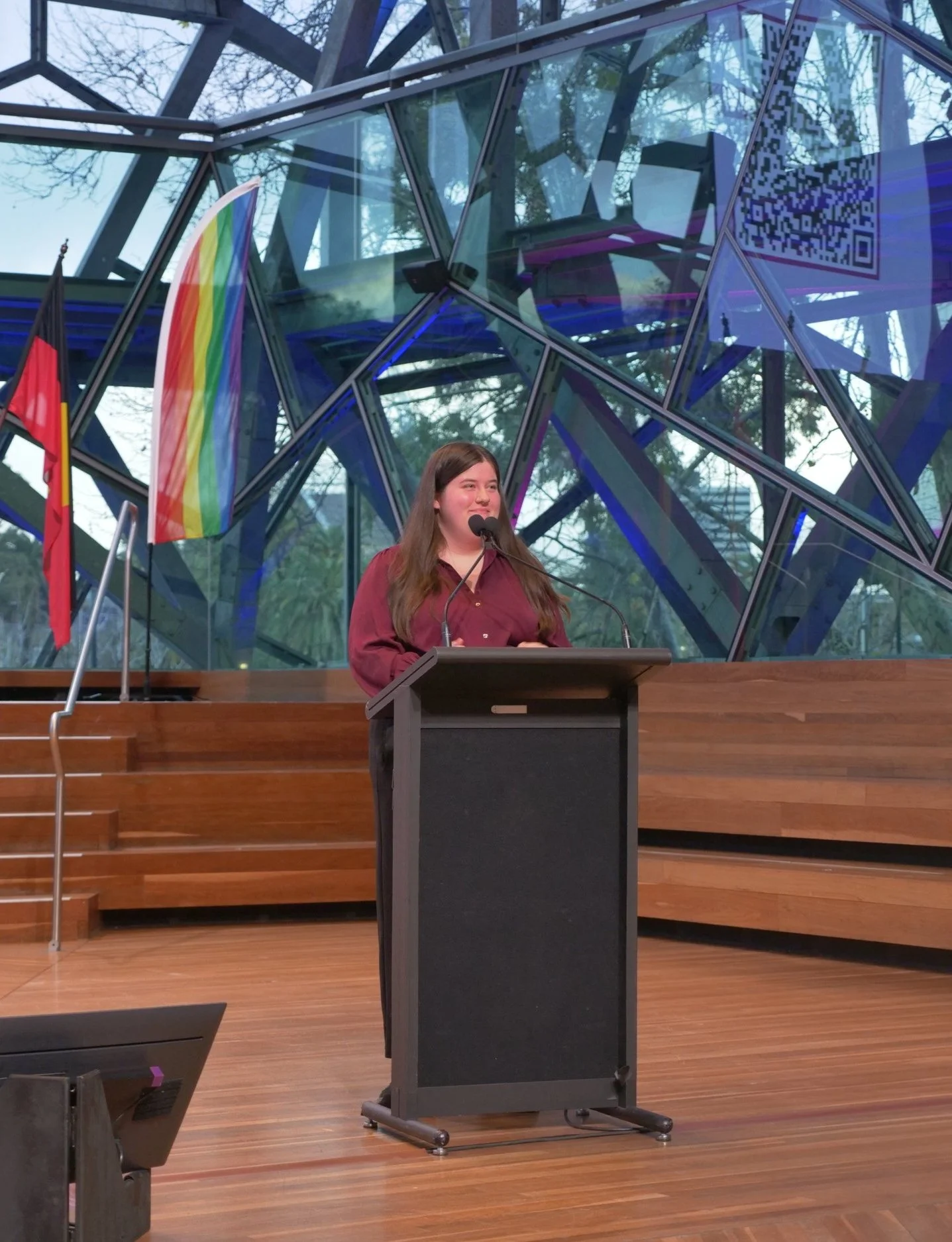 A young woman with long brown hair giving a speech at a podium in a modern building with glass walls, with a rainbow flag and another flag beside her.
