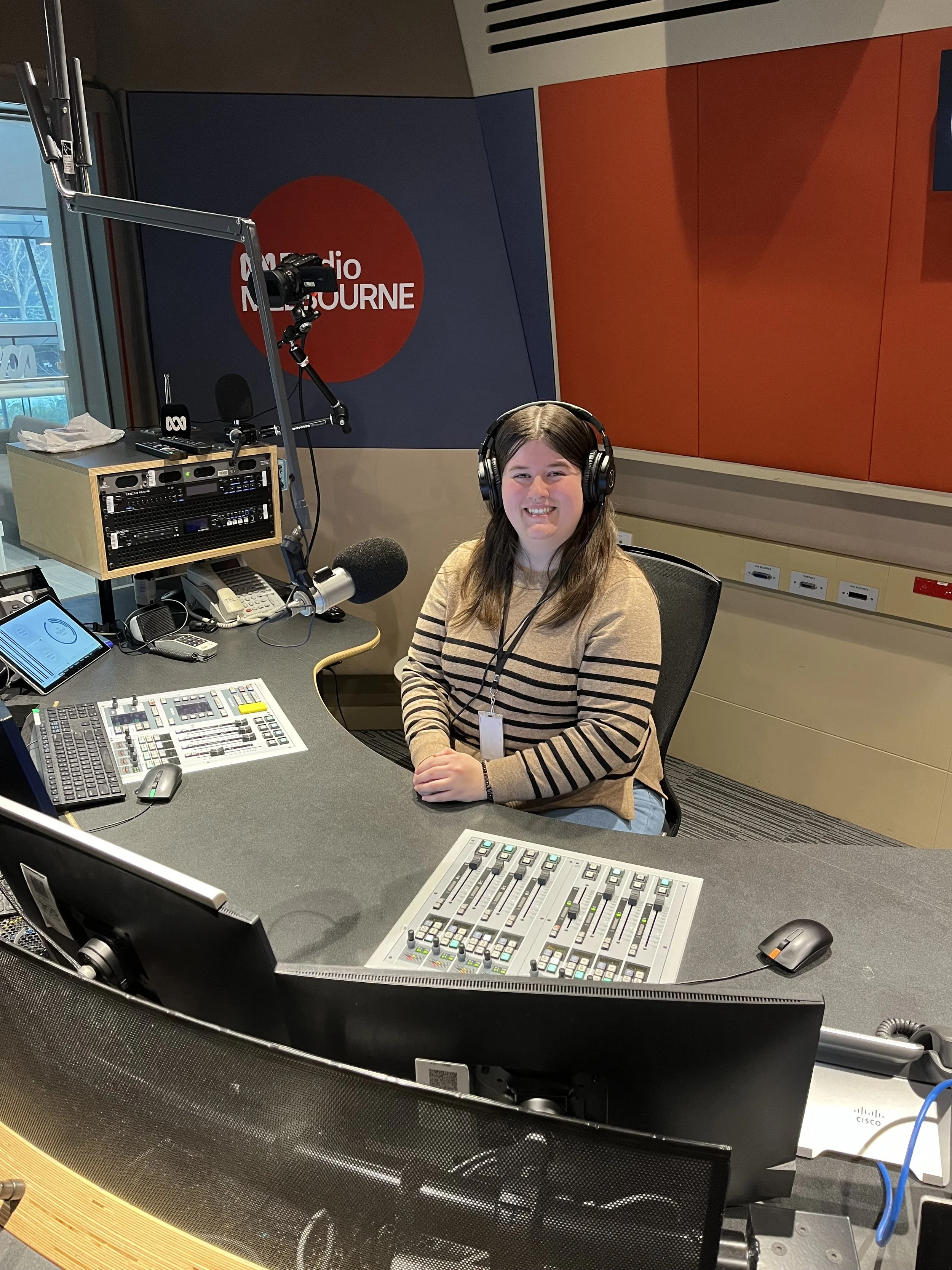 A woman with long brown hair wearing headphones, smiling, sitting in a radio studio with equipment including microphones, control panels, and computers, with the logo 'ABC Radio Melbourne' on the wall behind her.