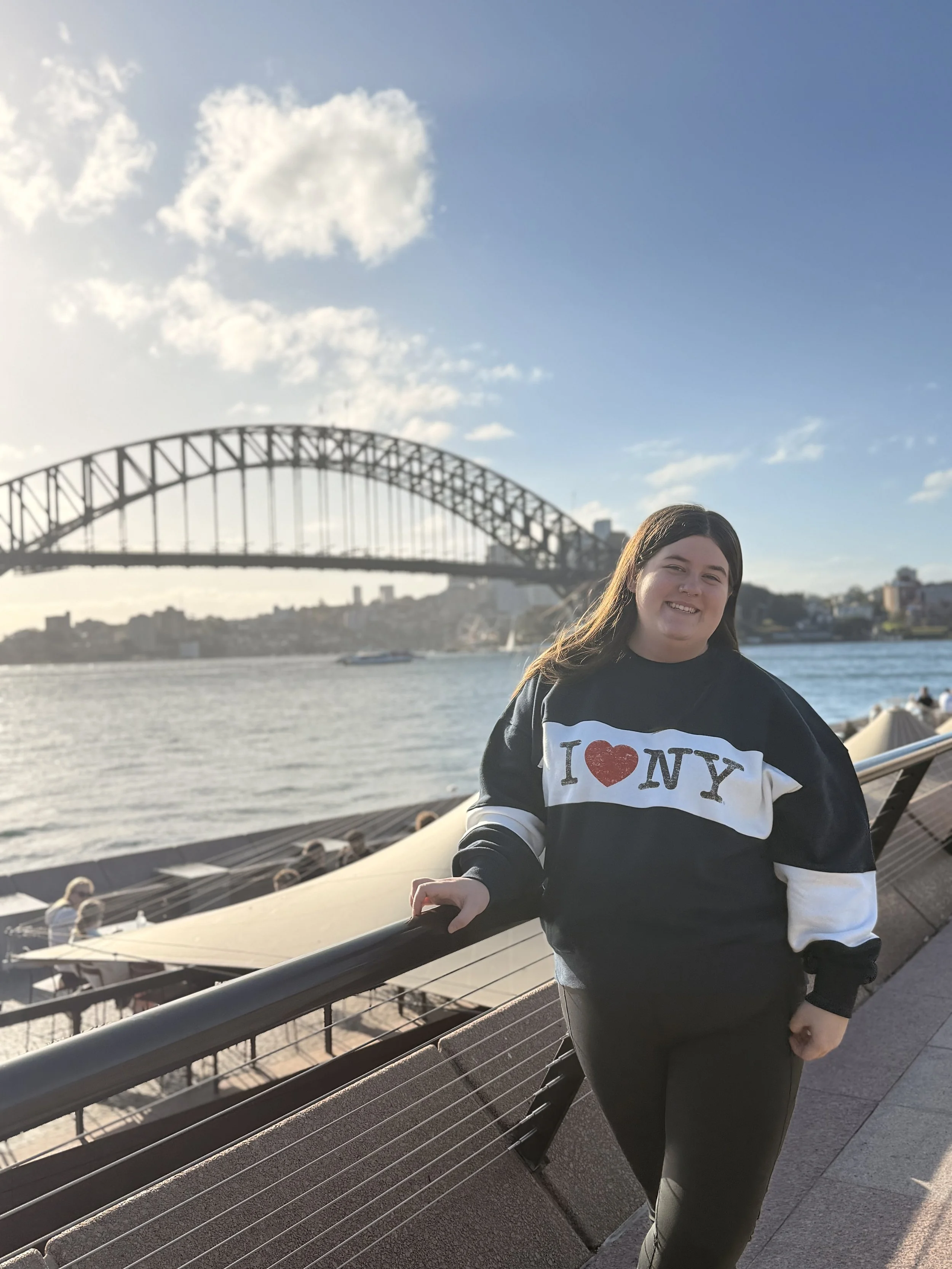 A young woman standing by the waterfront with the Sydney Harbour Bridge in the background, smiling at the camera, wearing a black and white sweatshirt that says "I ♥ NY".