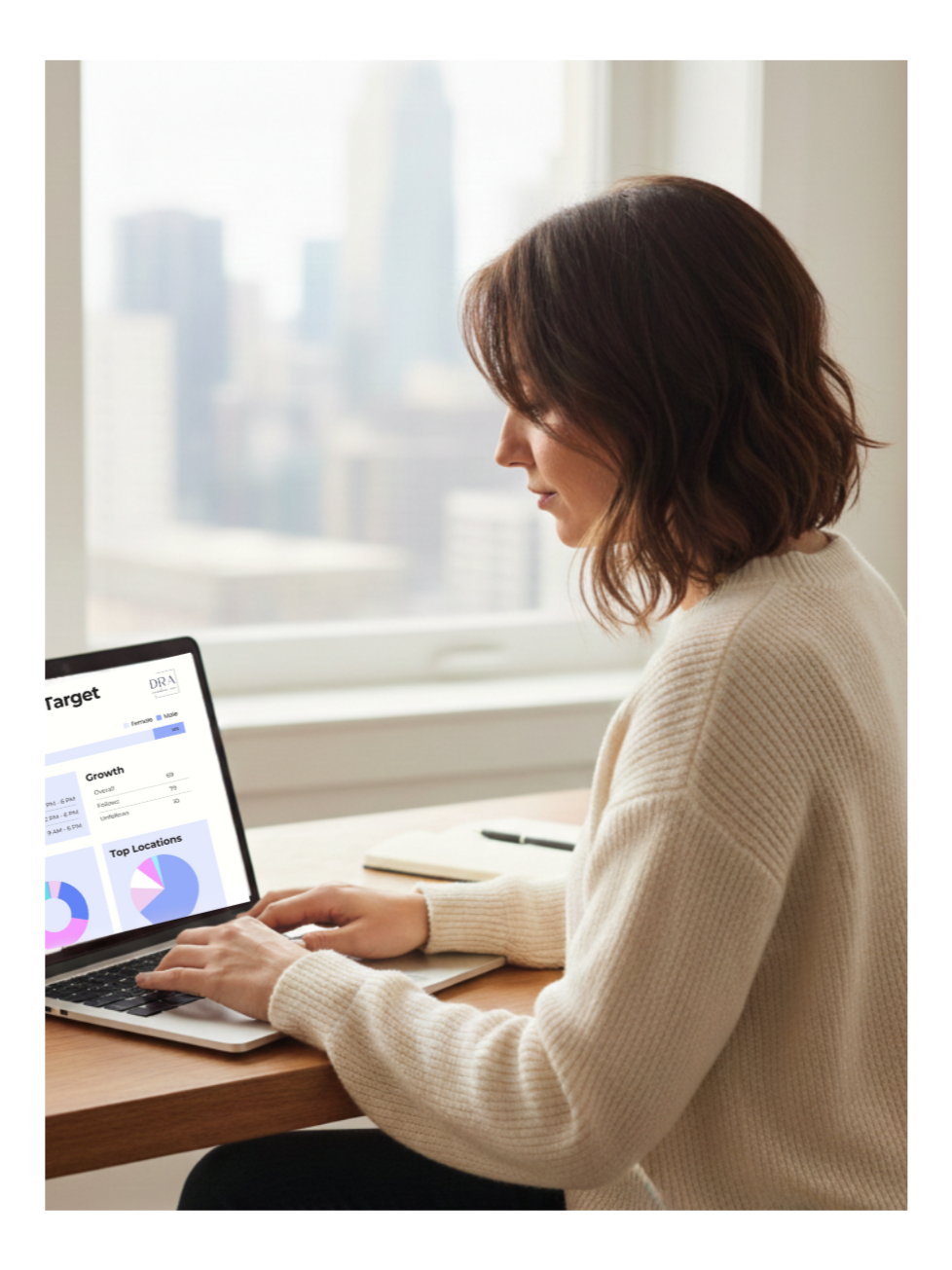 Woman with shoulder-length brown hair working on a laptop at a desk, with a cityscape visible through the window behind her.