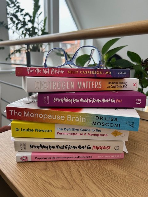 En stapel med böcker om perimenopaus och menopaus, på ett träbord vid ett fönster.