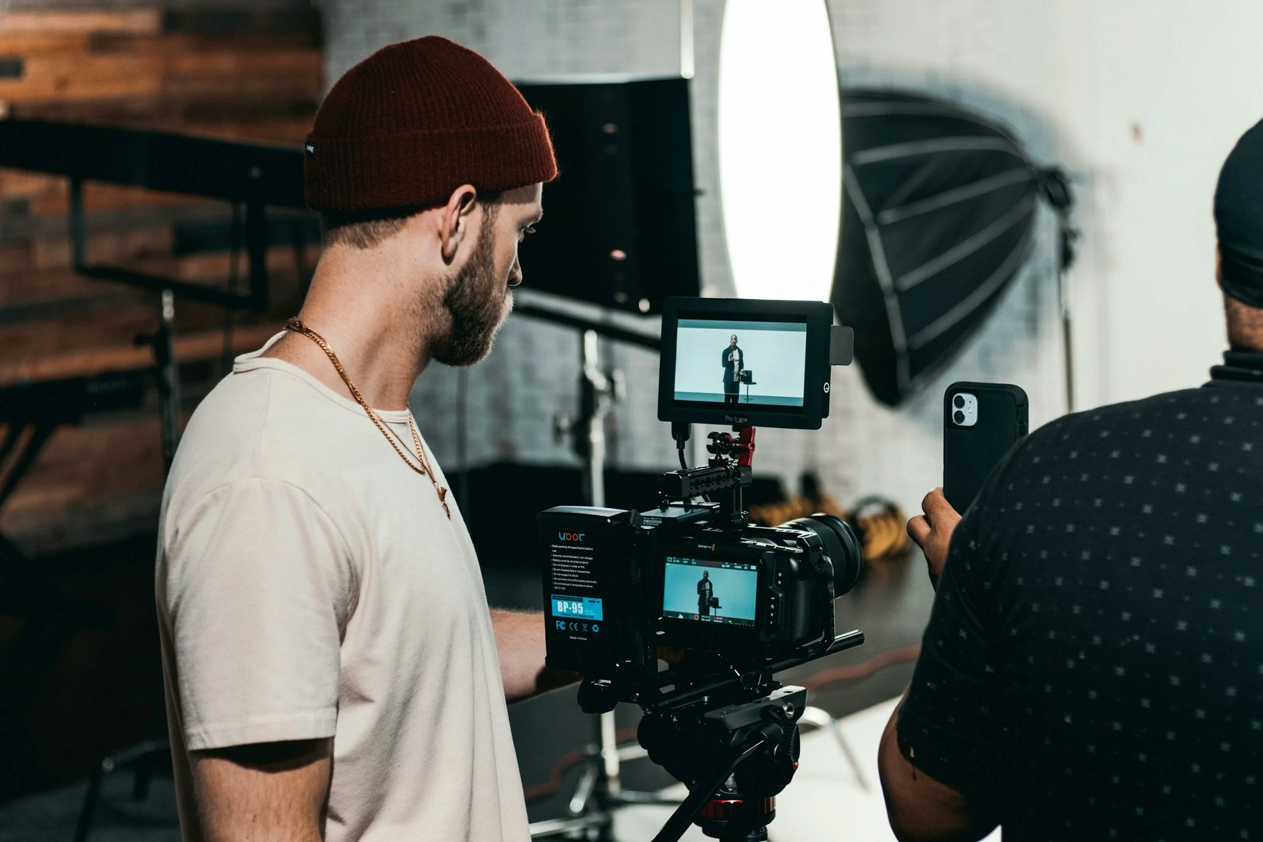 Two men working in a photography studio, one man is filming the other on a camera with a monitor, large softbox light in the background.