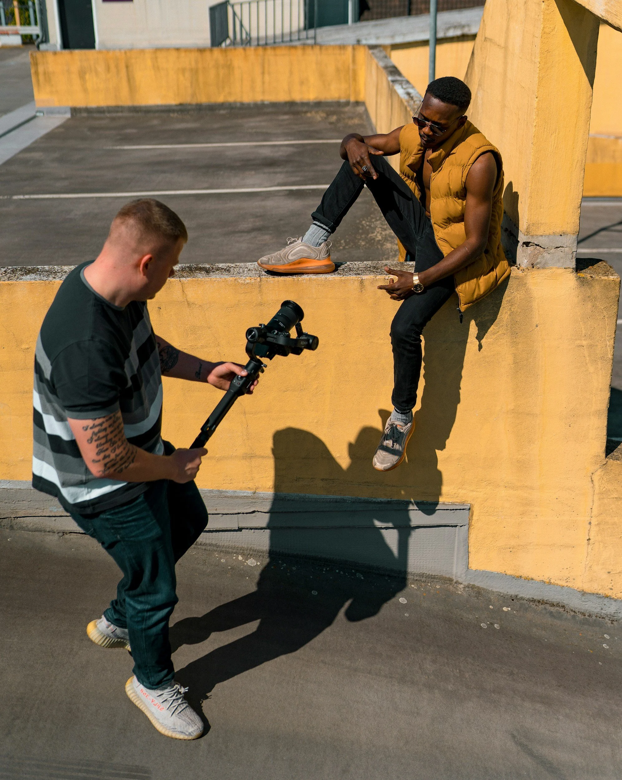 A man sitting on a yellow concrete wall nearby a person holding a camera stabilizer filming him. The man on the wall is wearing sunglasses, a brown vest, black pants, and sneakers, and is posing with one leg raised on the wall.