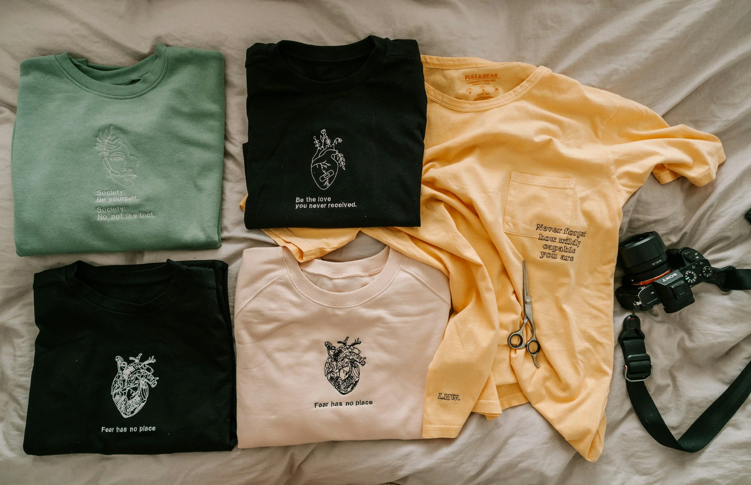 Six folded T-shirts in pastel and dark colors with printed designs and quotes, paired with a pair of scissors and a camera on a beige fabric surface.