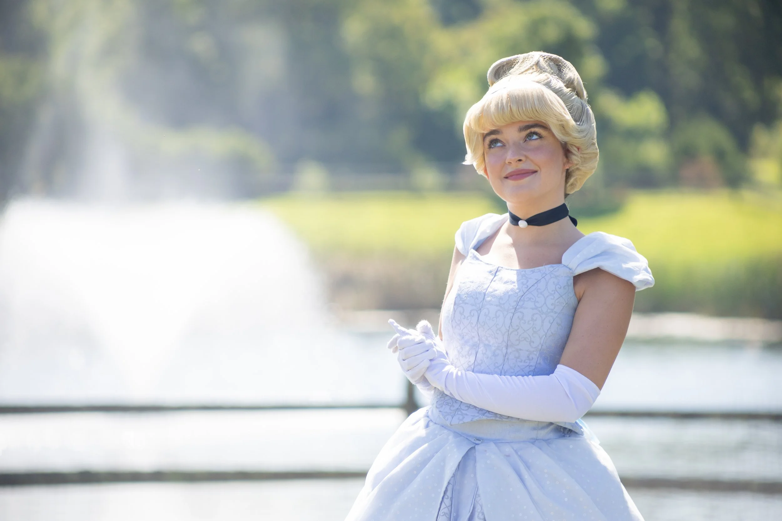 A woman dressed in a white vintage gown with elbow-length gloves, a choker, and a retro hairstyle, standing outdoors near water with a fountain in the background.