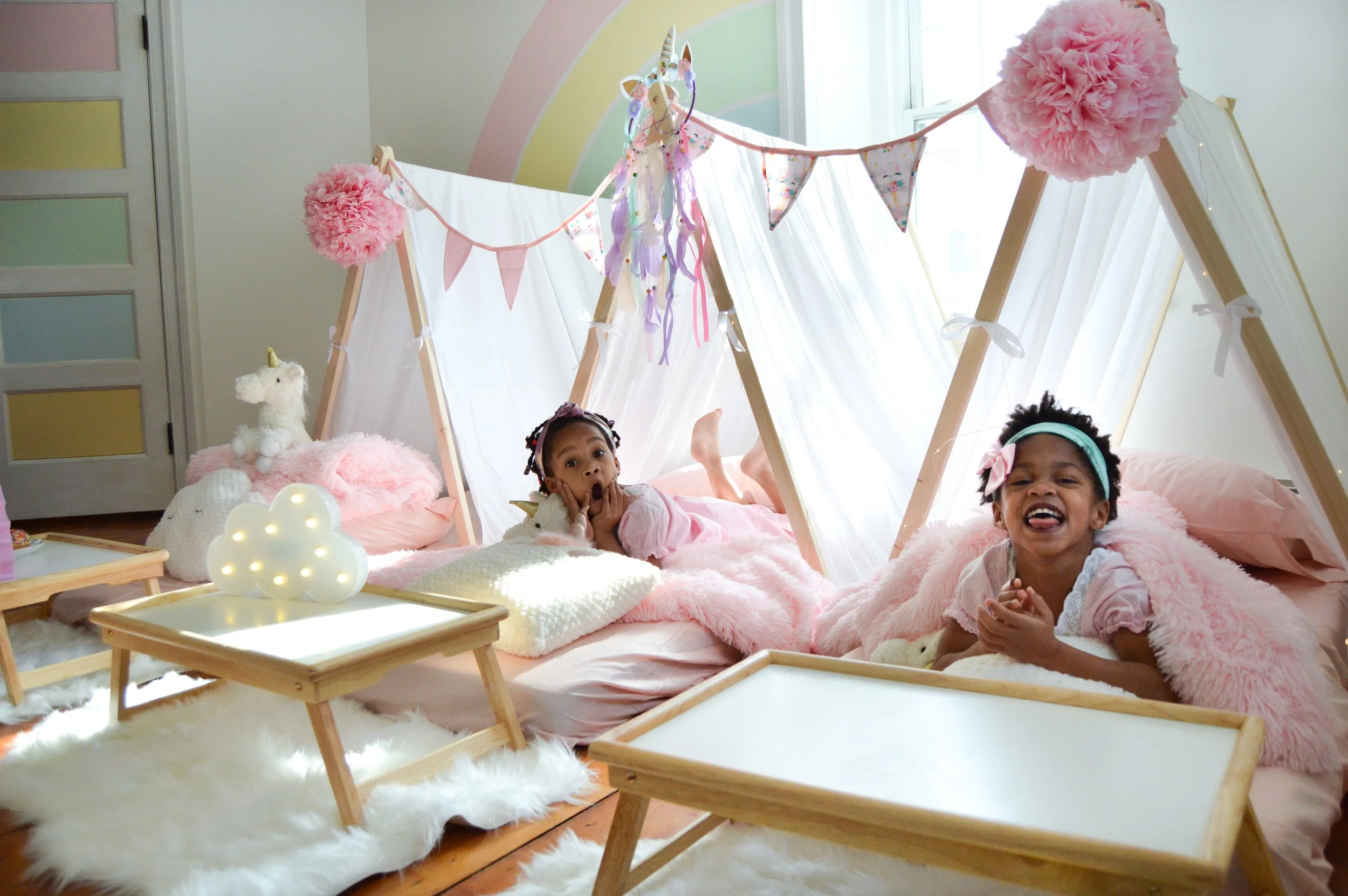 Two young girls are lying on pink beds under a playful pink canopy tent decorated with buntings and unicorn plush toys. The room has a pastel rainbow wall, and there are small wooden tables and white fluffy rugs around the beds.